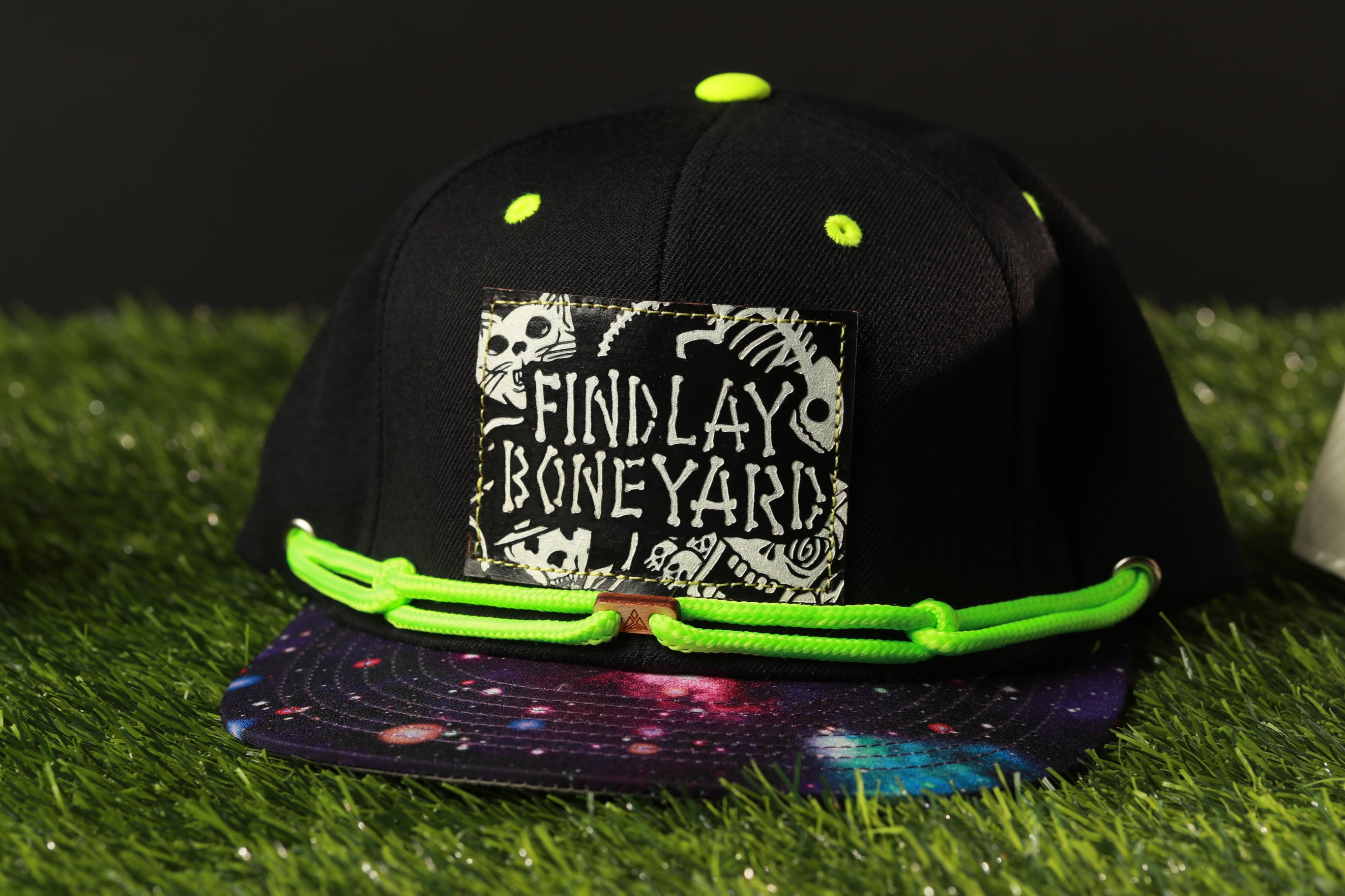 A black baseball cap with a colorful galaxy print and a patch that says "Findlay Boneyard" on it, placed on a grassy surface.