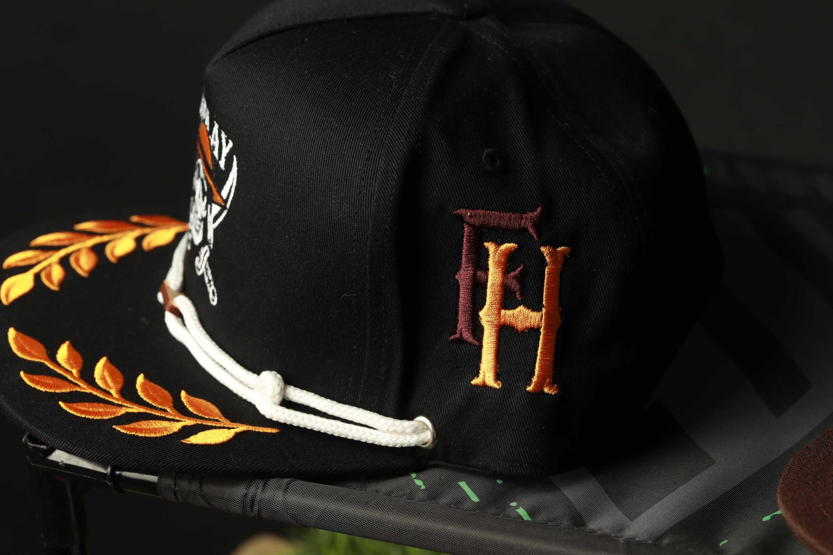 A black baseball cap with gold and orange embroidered details, including a monogrammed "F.H." logo.