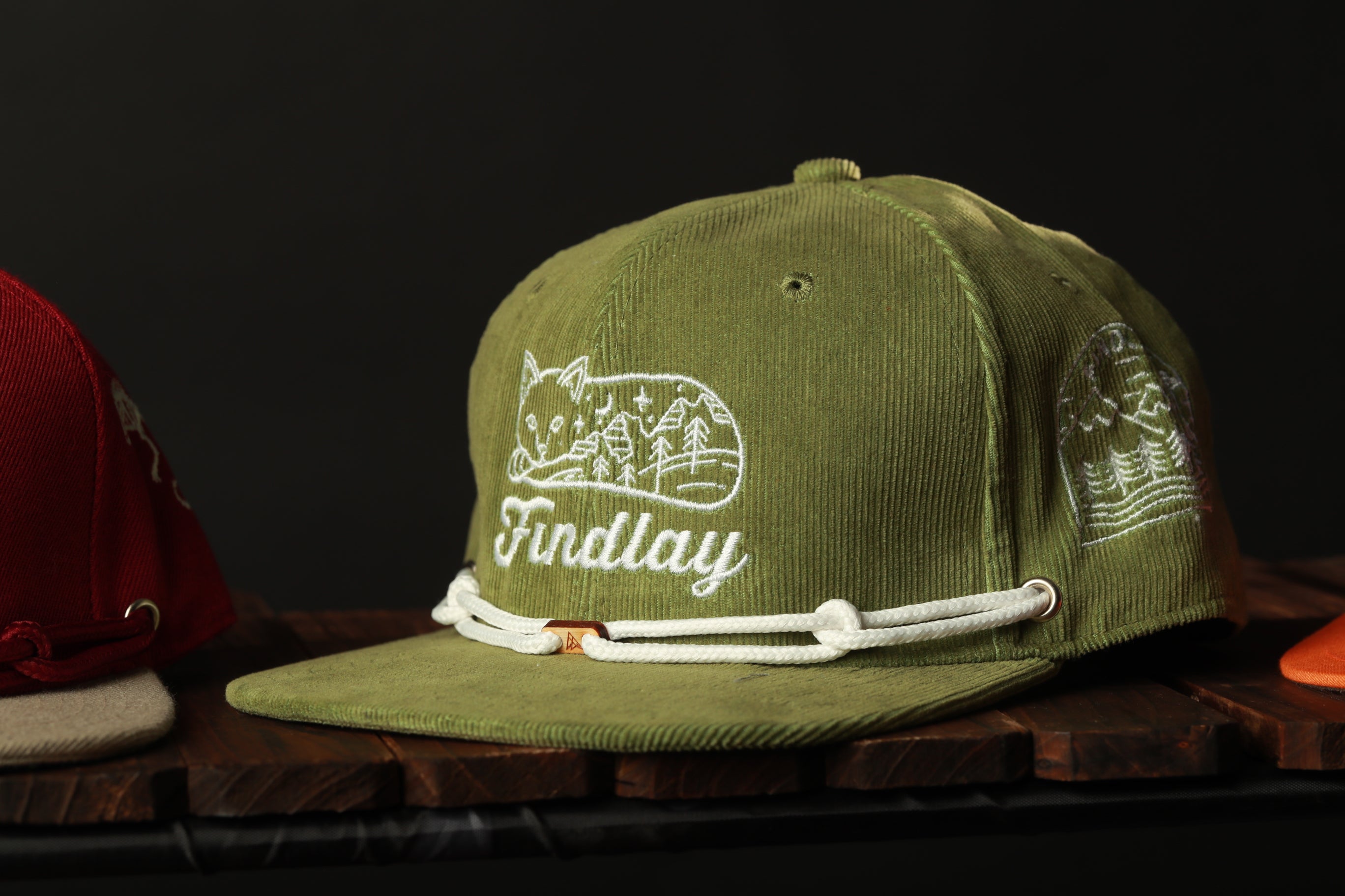A green baseball cap with a white embroidered design and the word "Findlay" printed on it, sitting on a wooden surface.