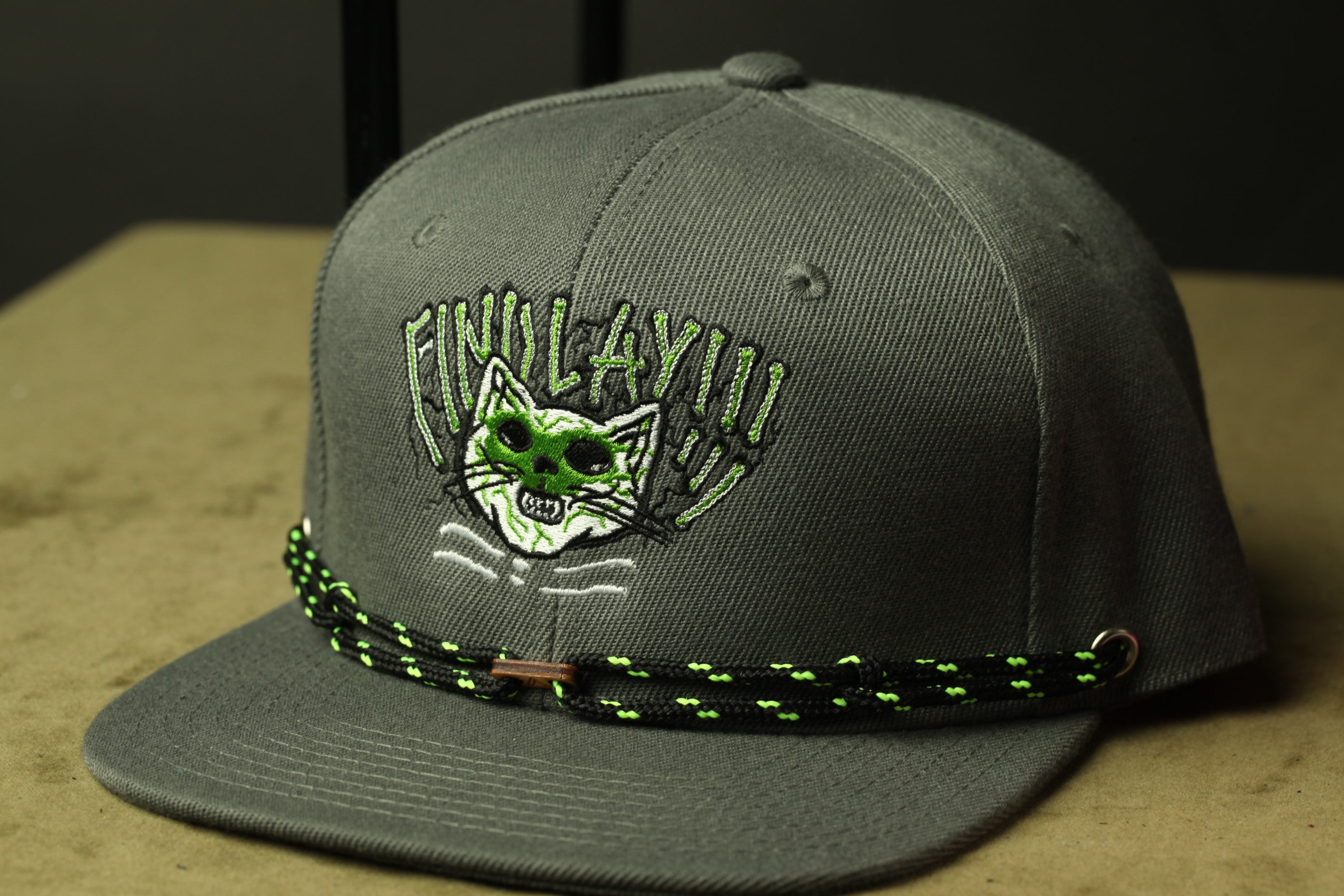 A gray baseball cap with a cat face embroidered on it, wearing a green and black braided strap.