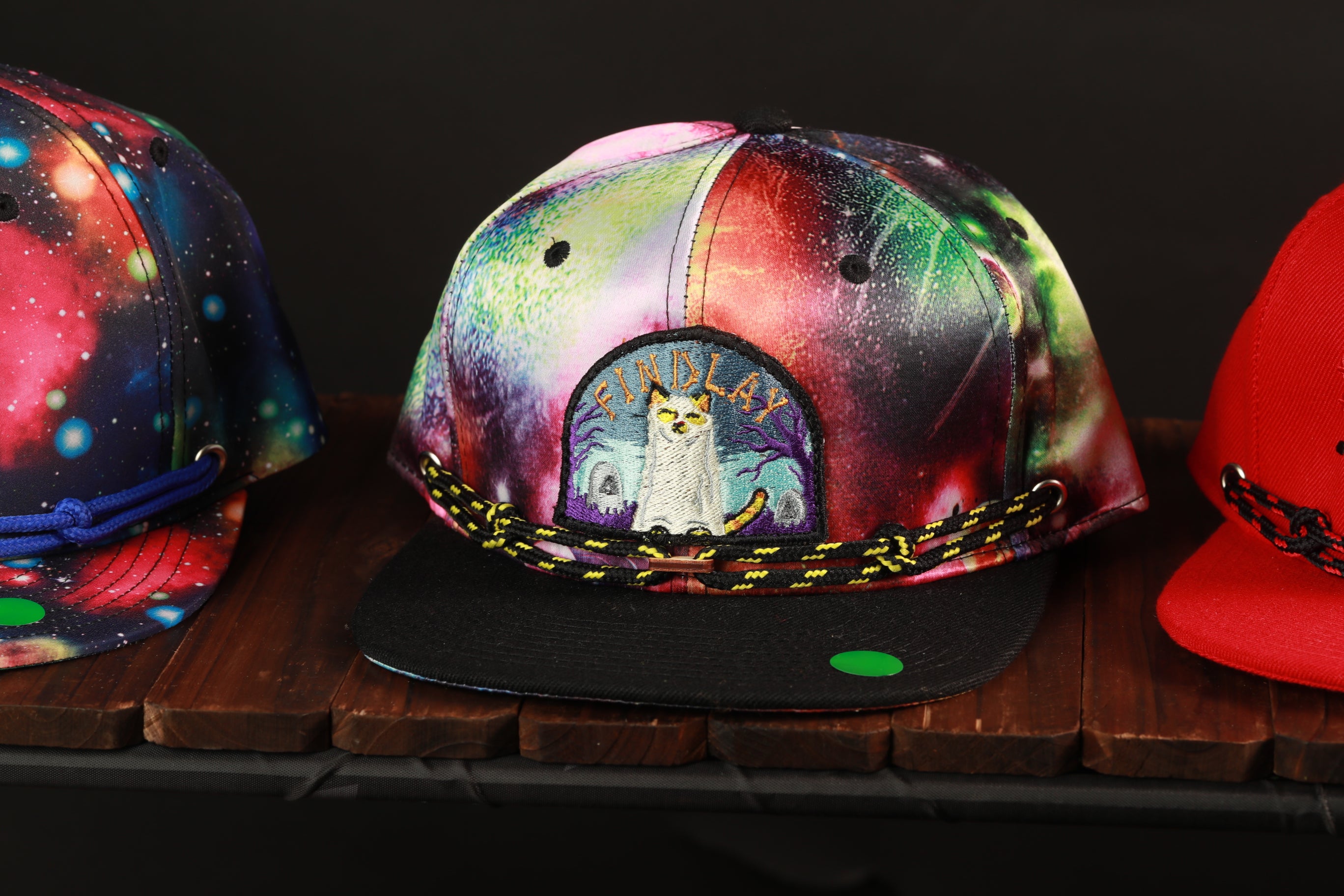 A colorful, patterned baseball cap with a cat design on it, displayed on a wooden surface.