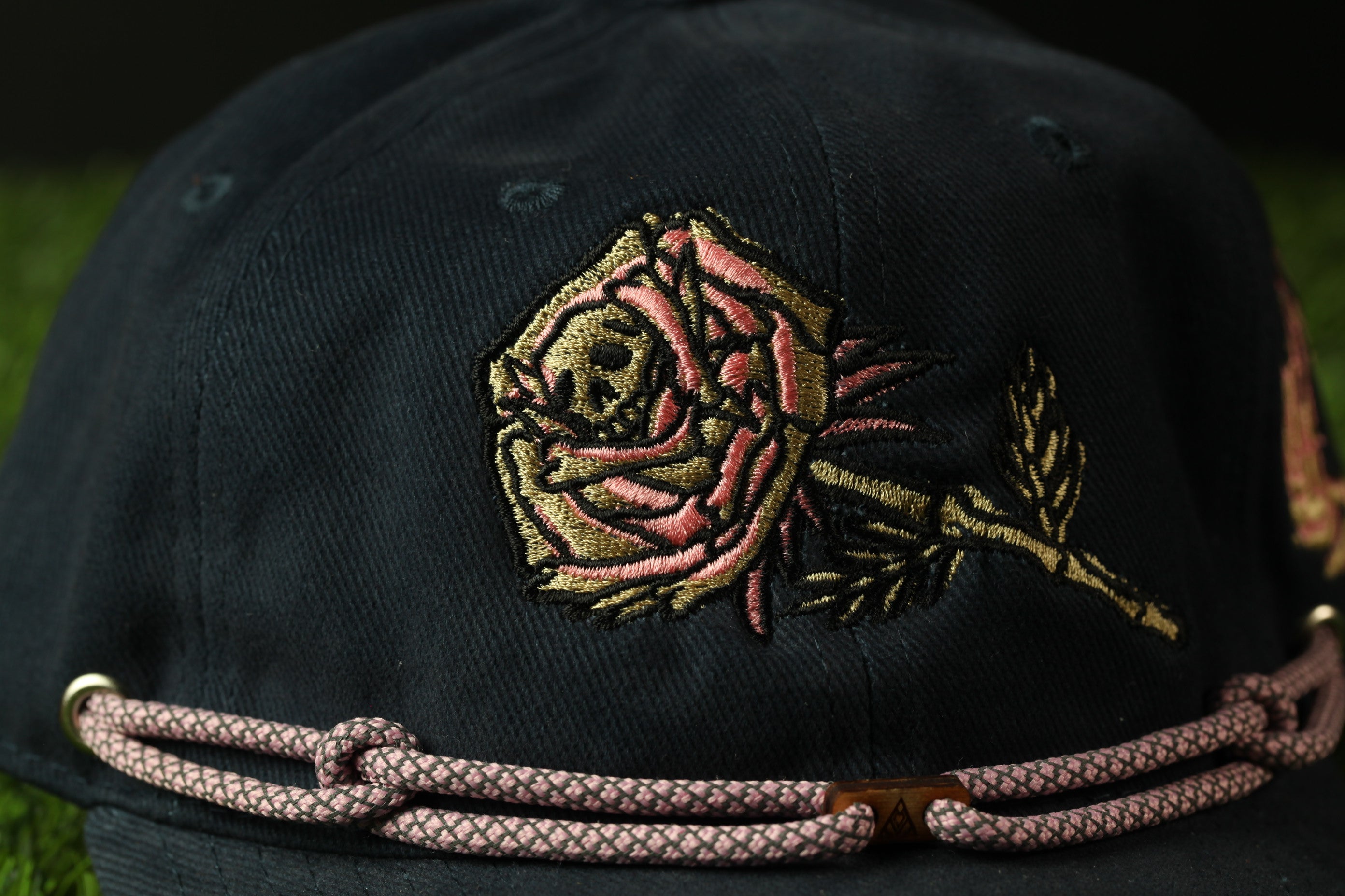 A close-up view of a black baseball cap with a pink and gold embroidered rose design on the front.