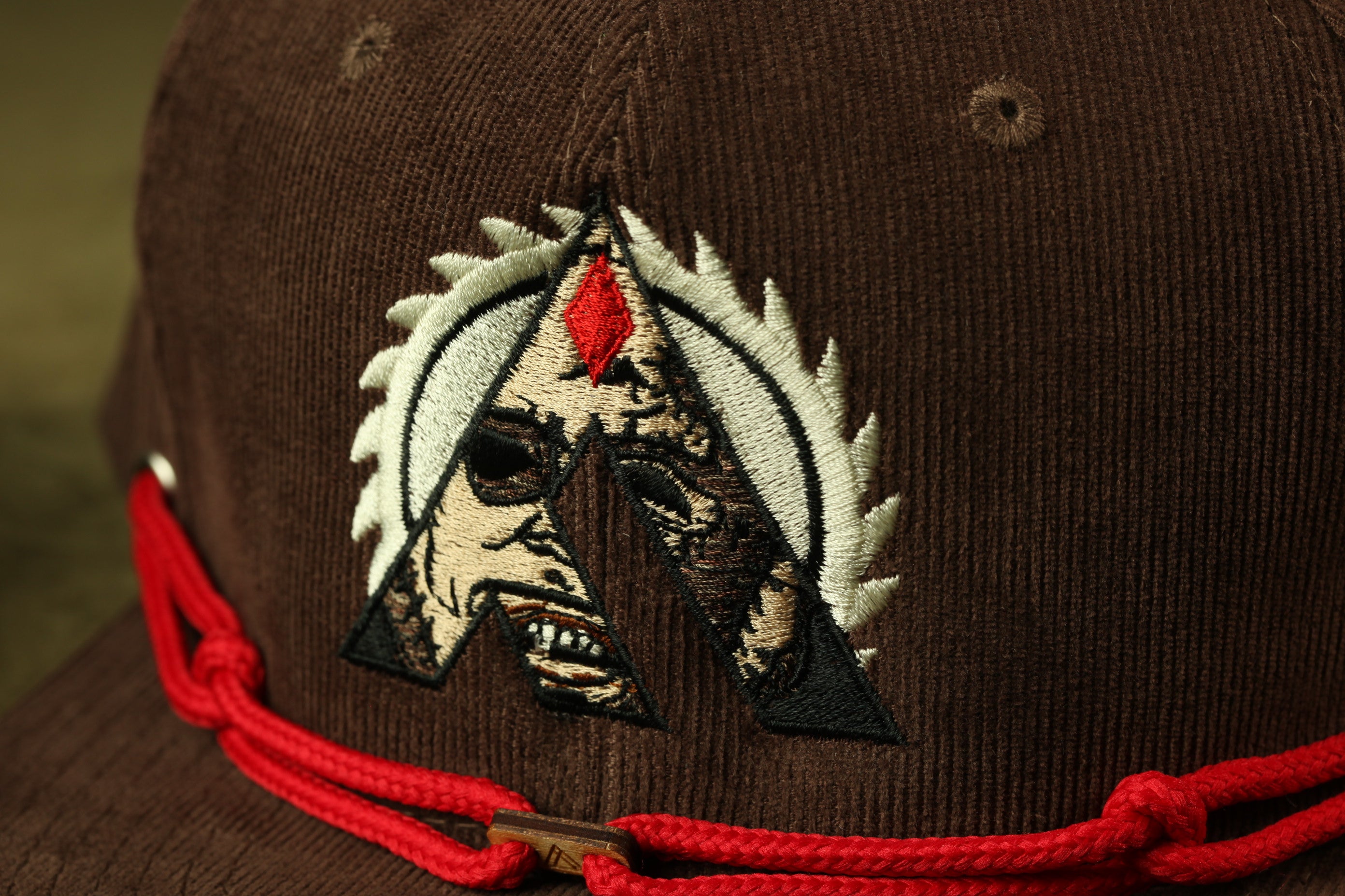 a close-up view of a brown baseball cap with a red string around the brim. The cap has an embroidered logo on the front, which appears to be a stylized letter 'A' with a red gem or stone in the center.