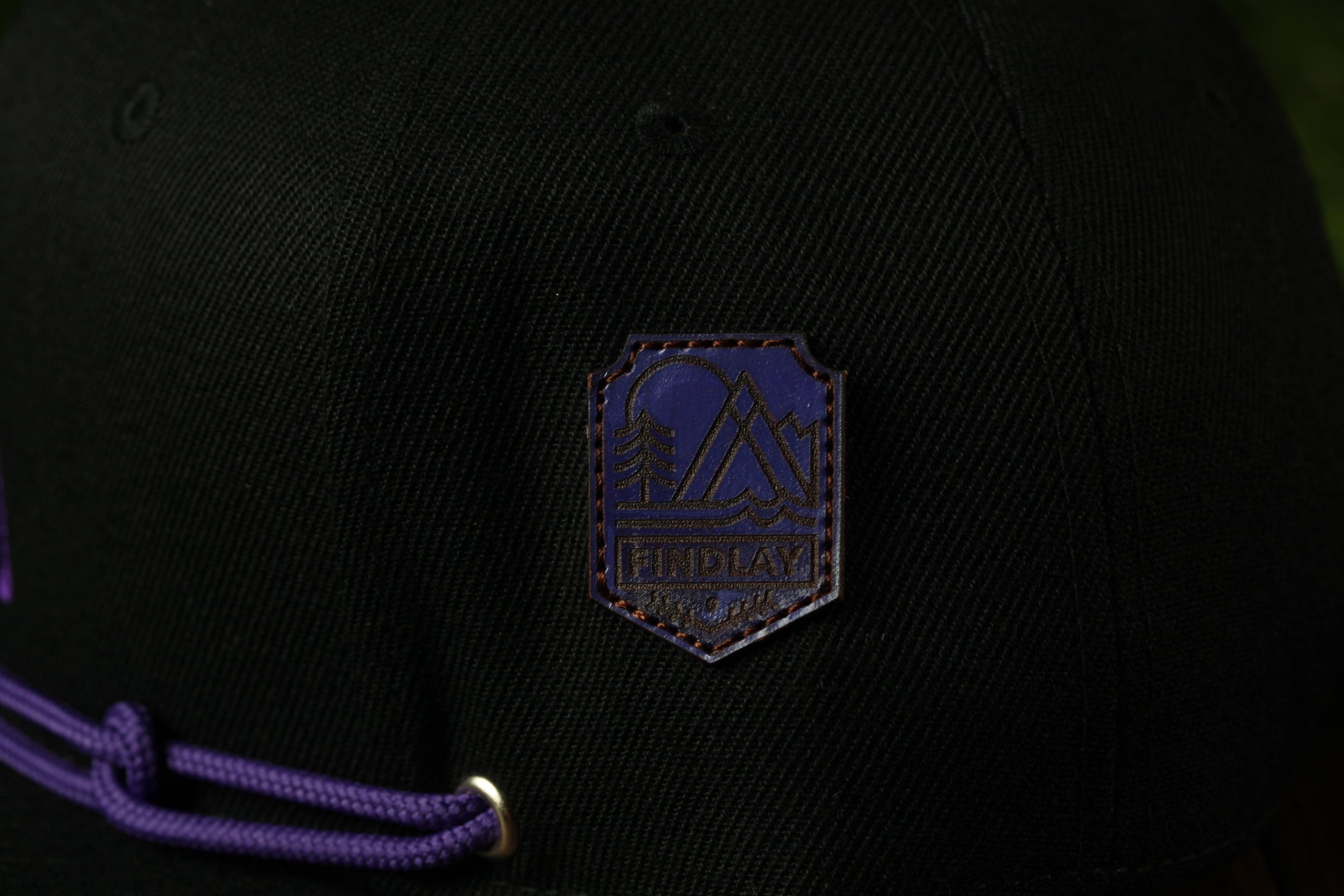 A close-up view of a black baseball cap with a purple logo on the front.