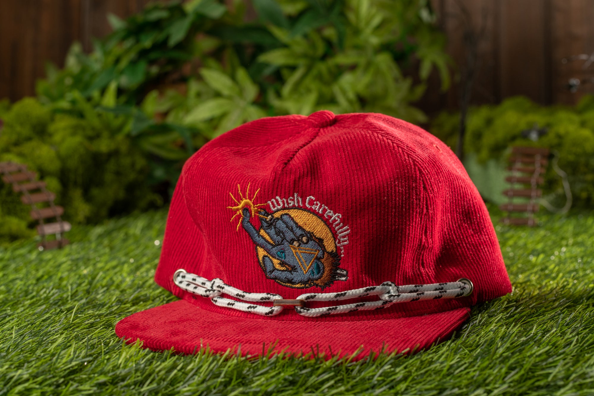 A red baseball cap with a graphic design sits on a grassy surface, with a wooden fence and greenery in the background.