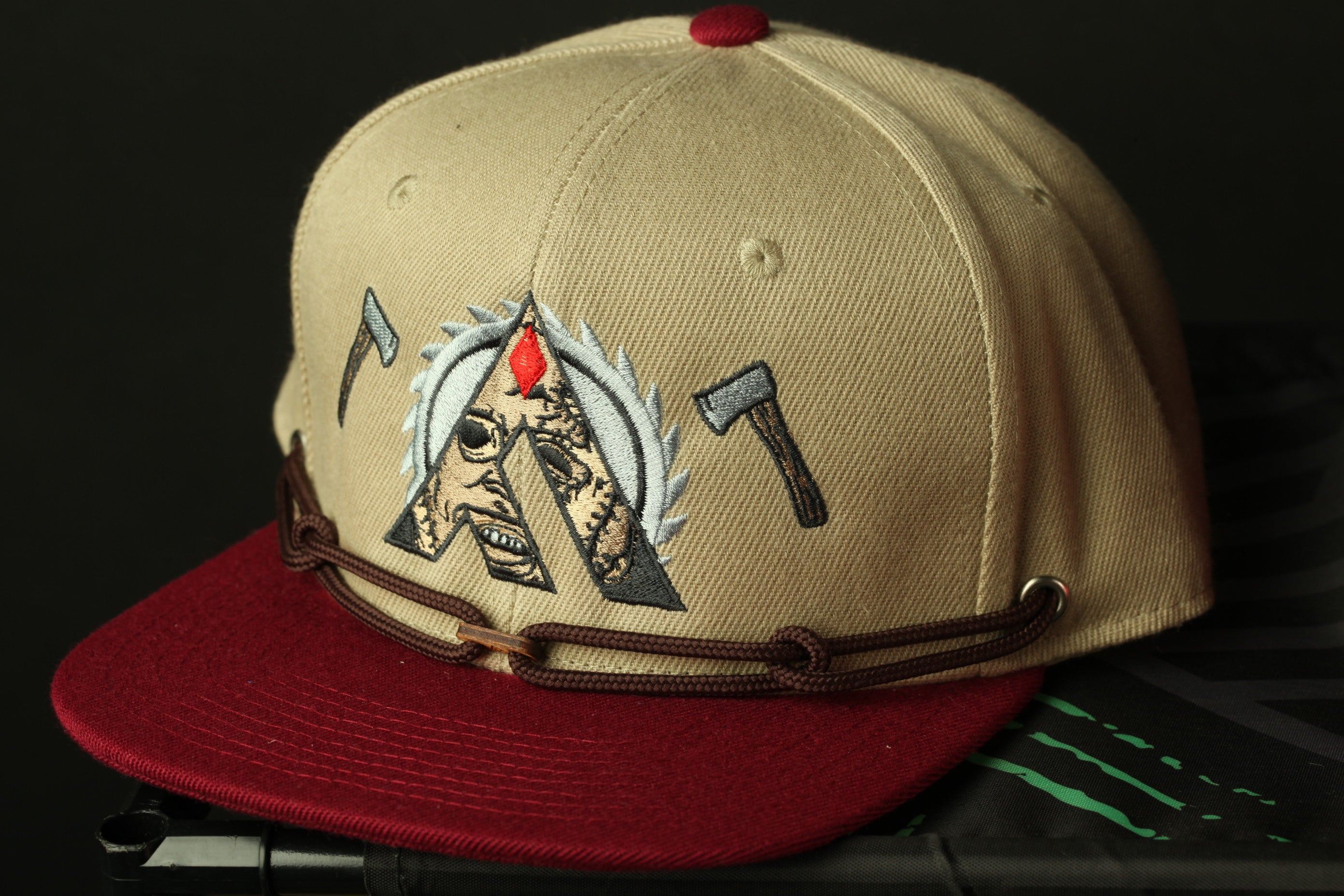 A tan baseball cap with a red brim and brown leather strap, featuring a graphic design with a skull and crossbones symbol.