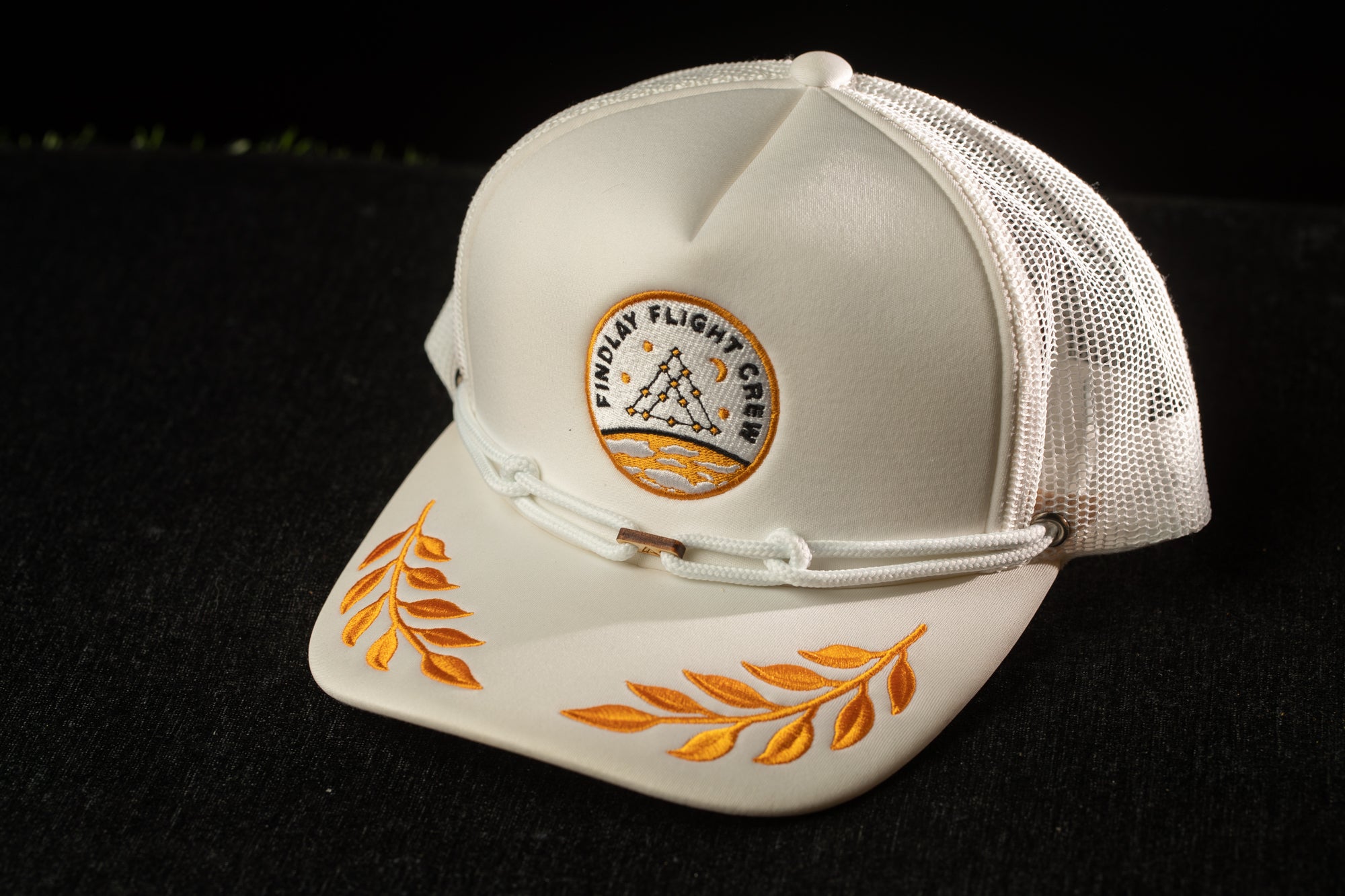 A white baseball cap with a mesh back and a circular logo on the front, featuring a design with leaves and a ship.