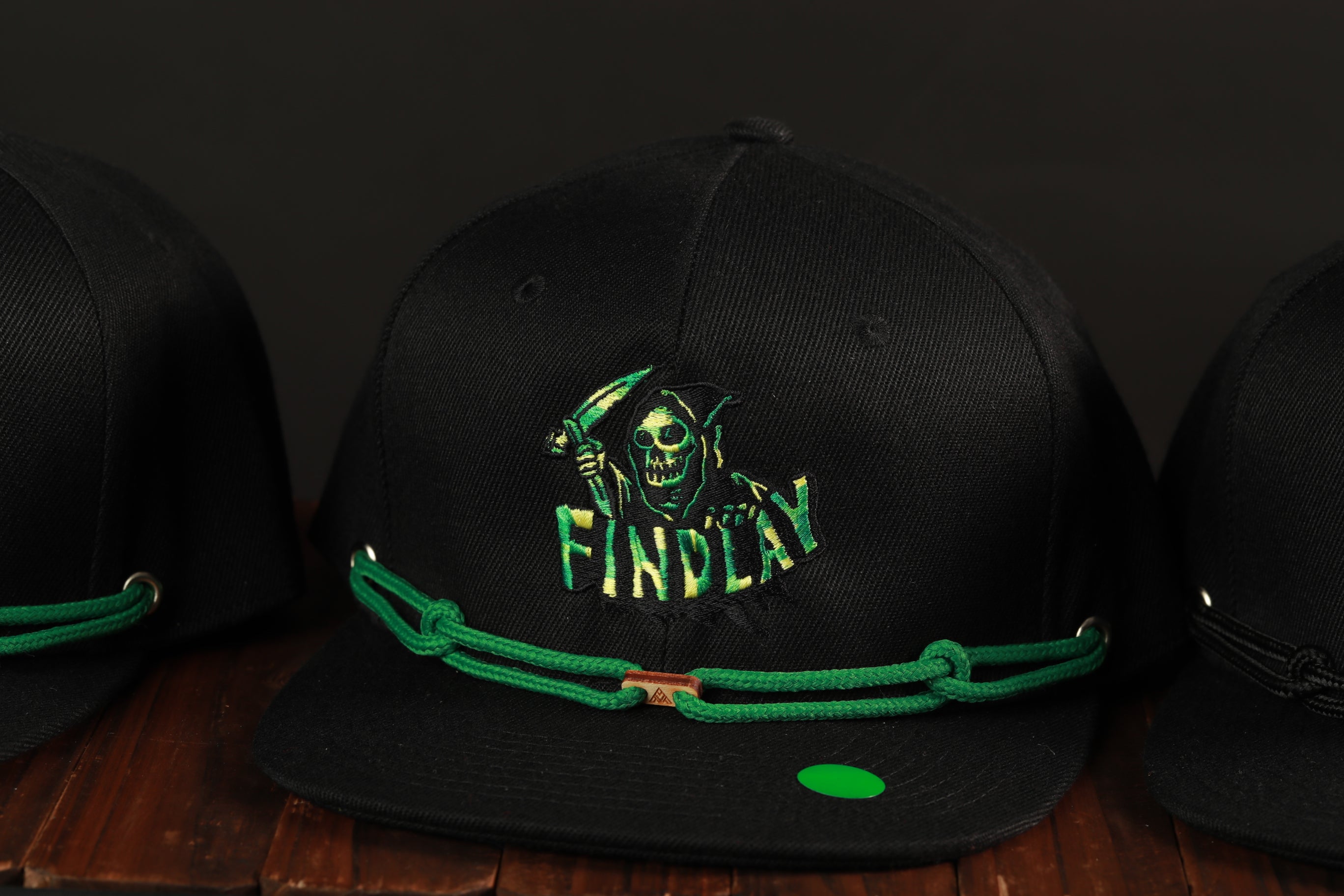 A black baseball cap with a green chain and a skull logo on it, displayed on a wooden surface.
