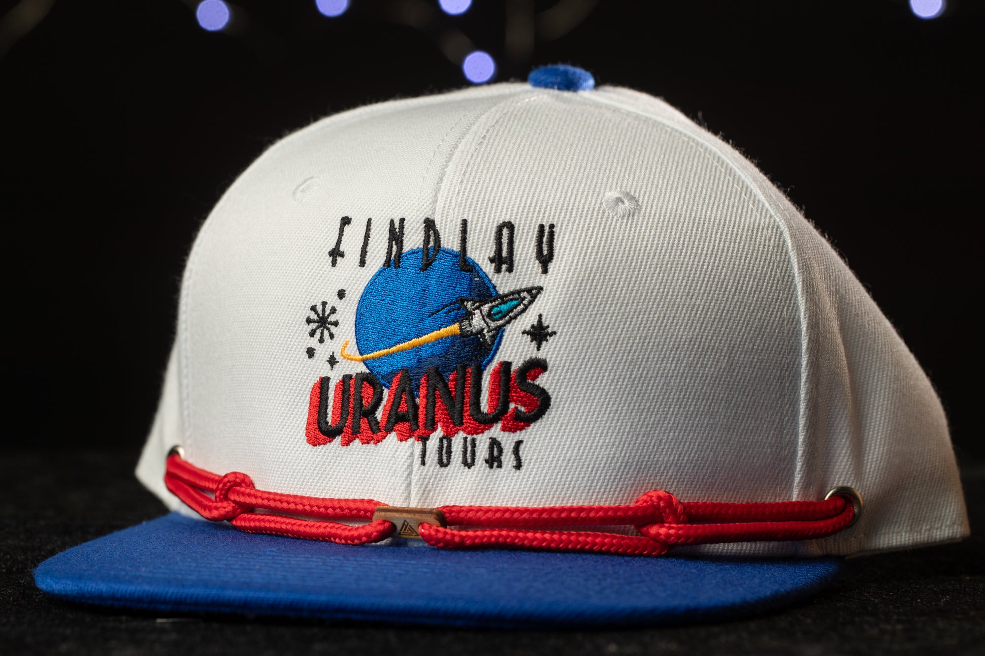A white baseball cap with a blue brim and red string, featuring a logo for "Findlay Uranus Tours".