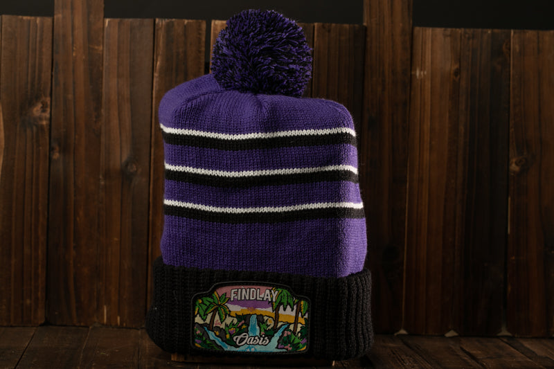 A purple and black striped beanie with a pom pom on top, placed on a wooden surface against a wooden background.