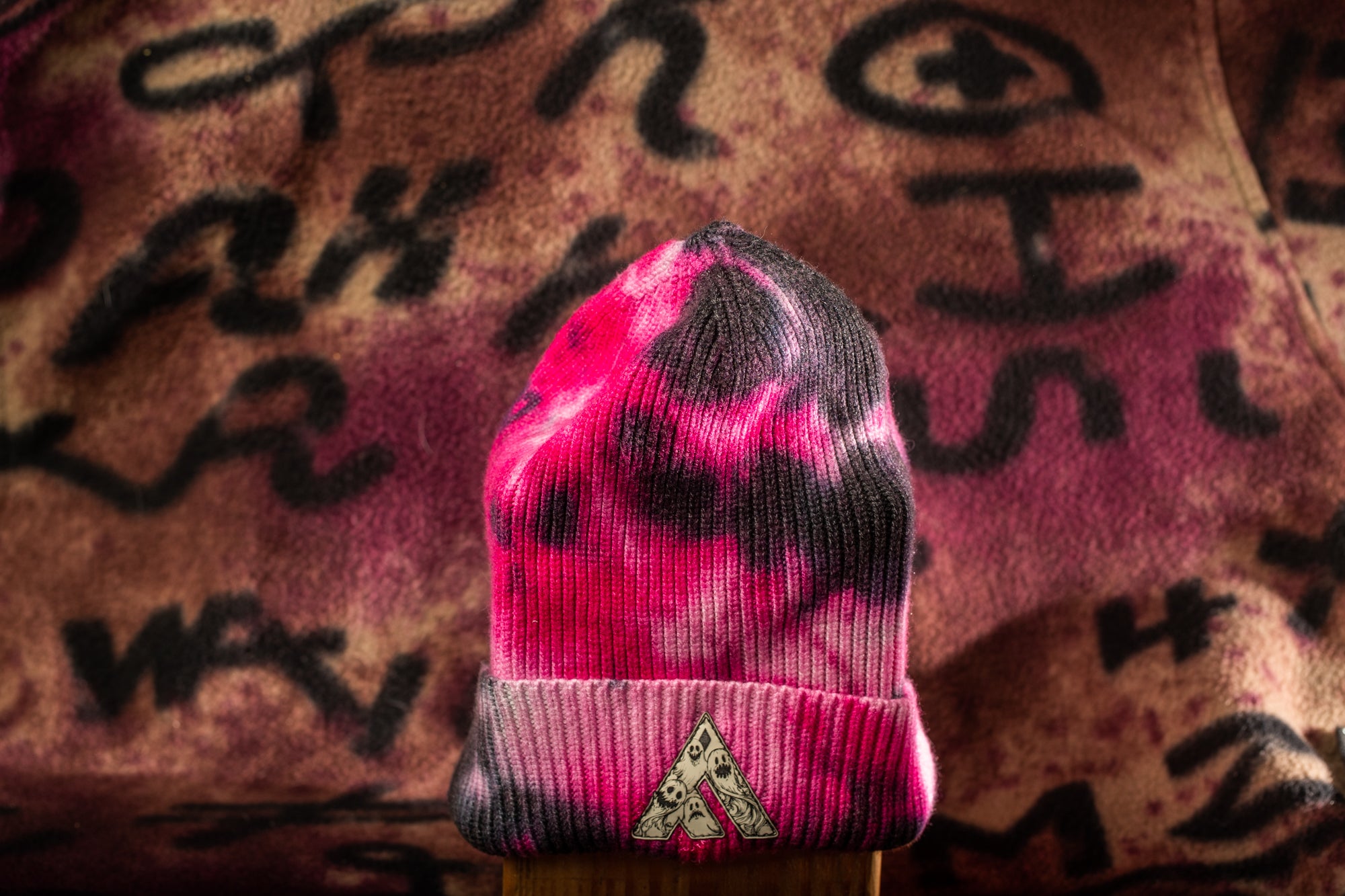 A tie-dye beanie with a pink and black pattern is displayed against a background with graffiti-style text.