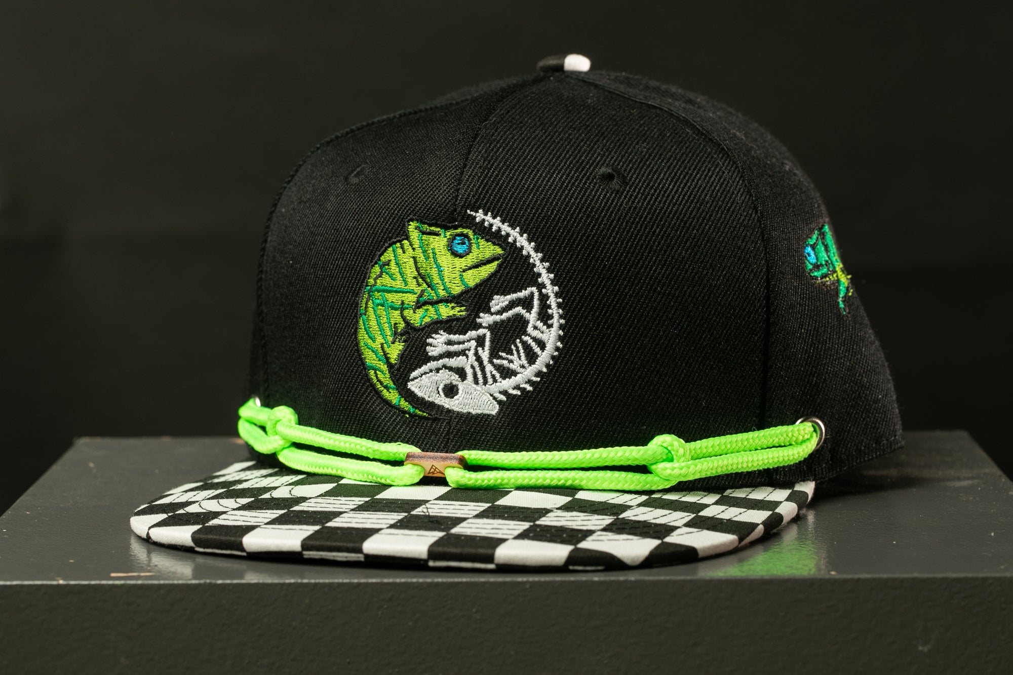 A black baseball cap with a green and white checkered pattern and a graphic of a chameleon on the front.