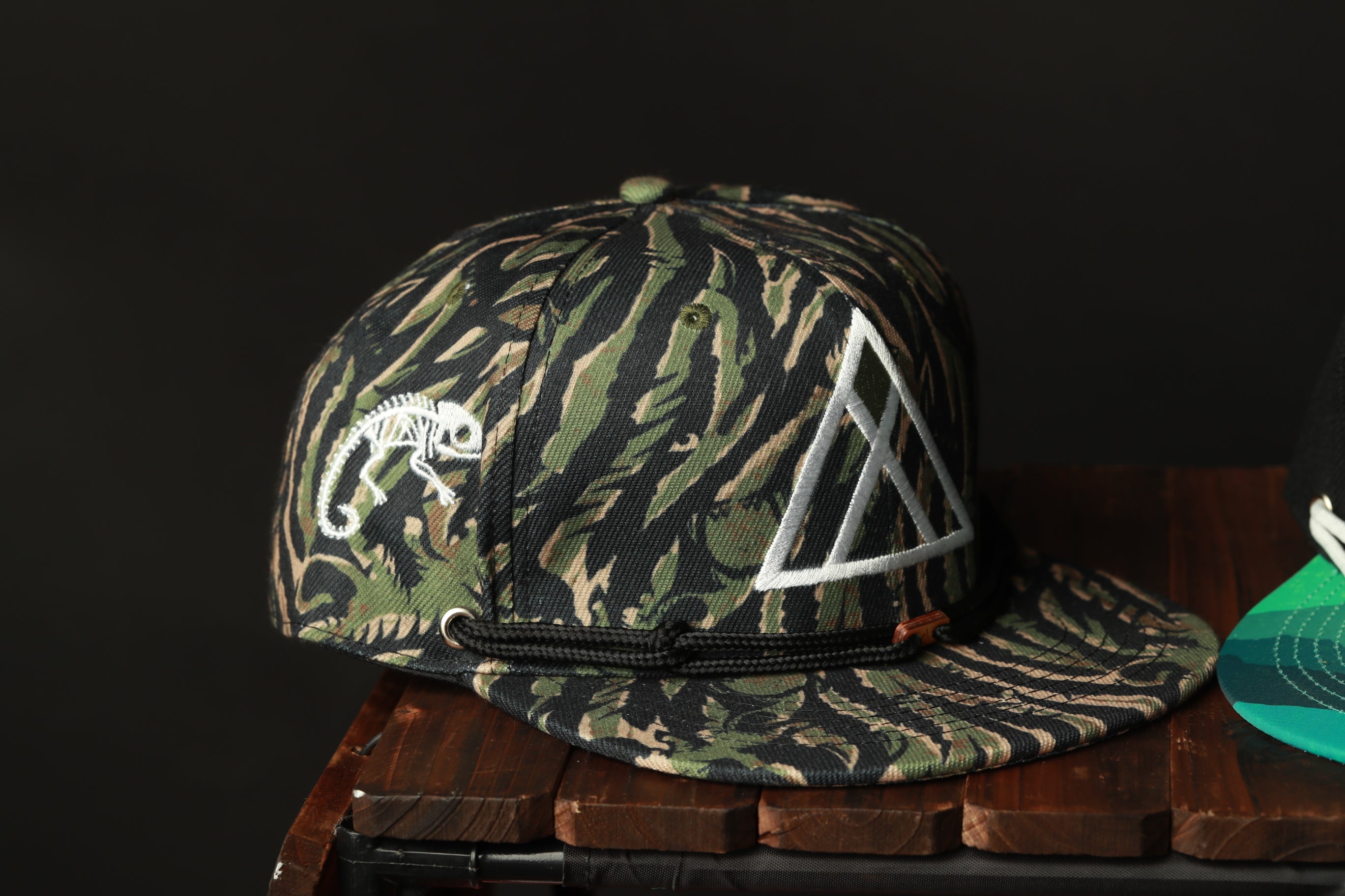 A camouflage baseball cap with a white triangle design on the front, placed on a wooden surface.