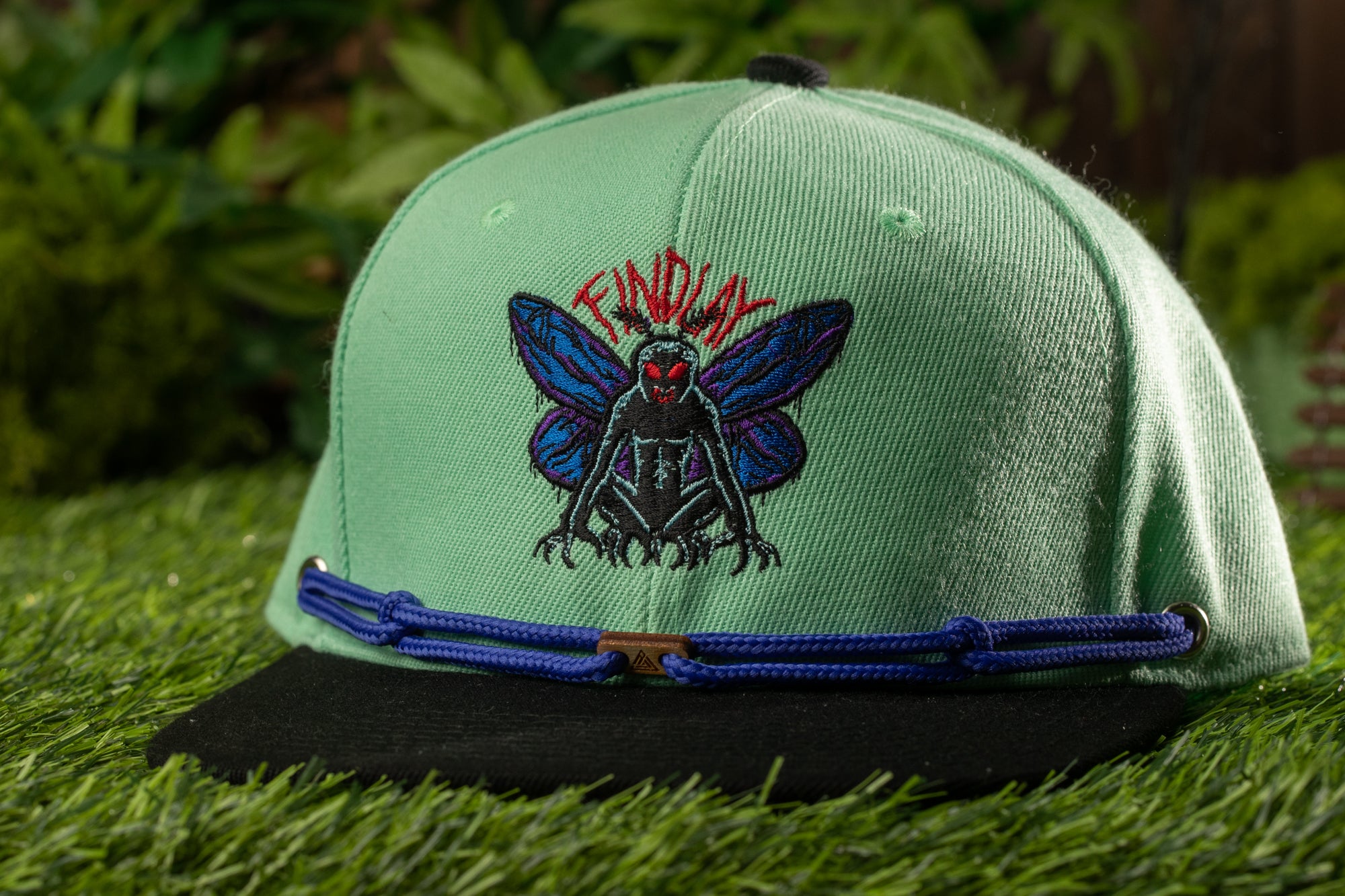 A green baseball cap with a blue and black design featuring a large, colorful insect-like creature on the front.