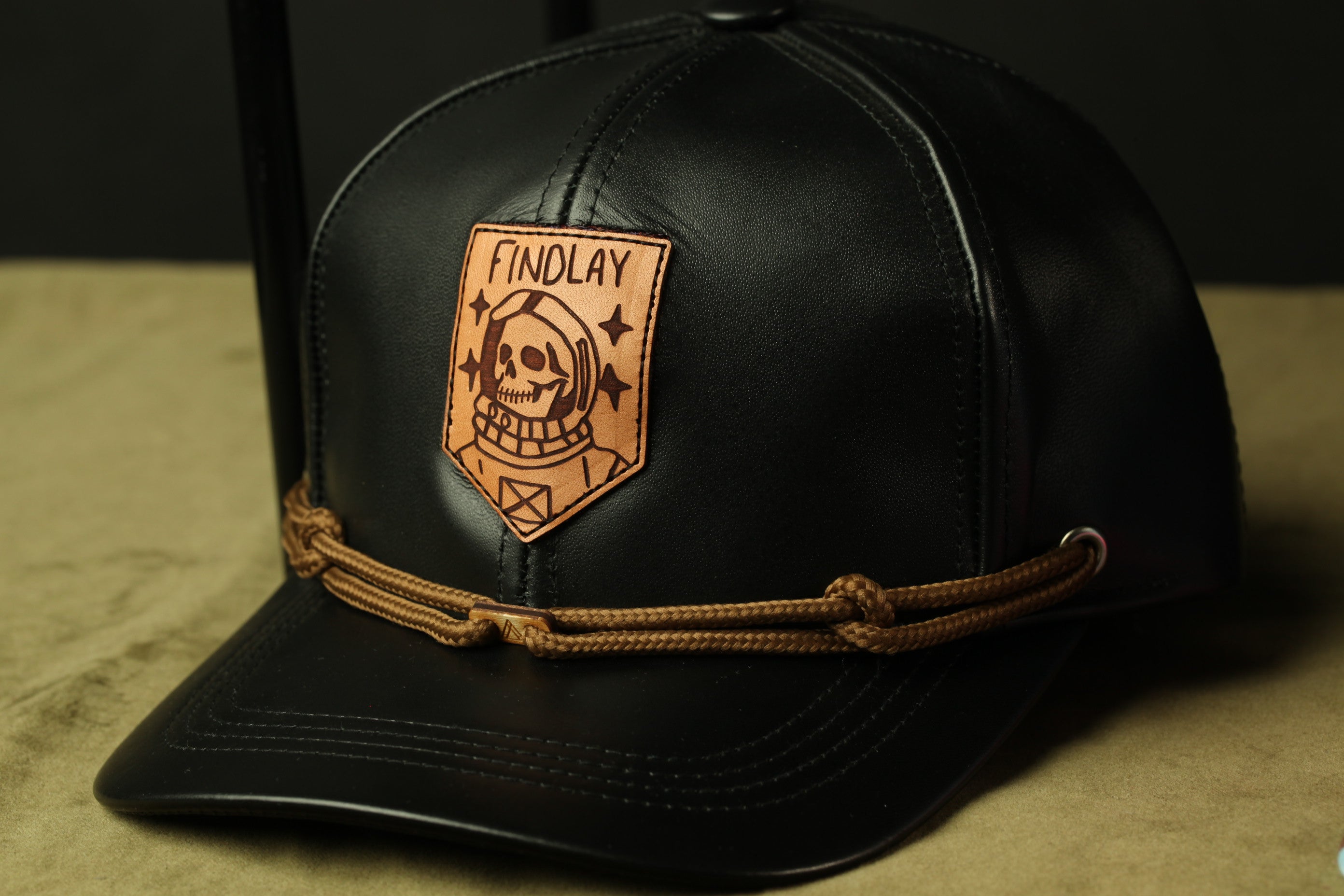 A black baseball cap with a leather patch featuring a skull and the word "FINDOLAY" printed on it.
