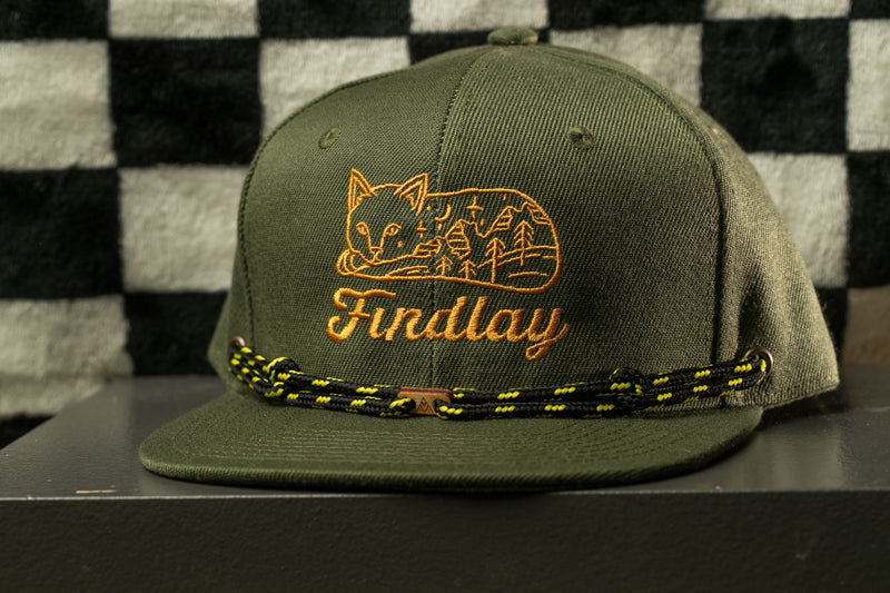 A green baseball cap with a cat design and the word "Findlay" printed on it, sitting on a gray surface against a black and white checkered background.