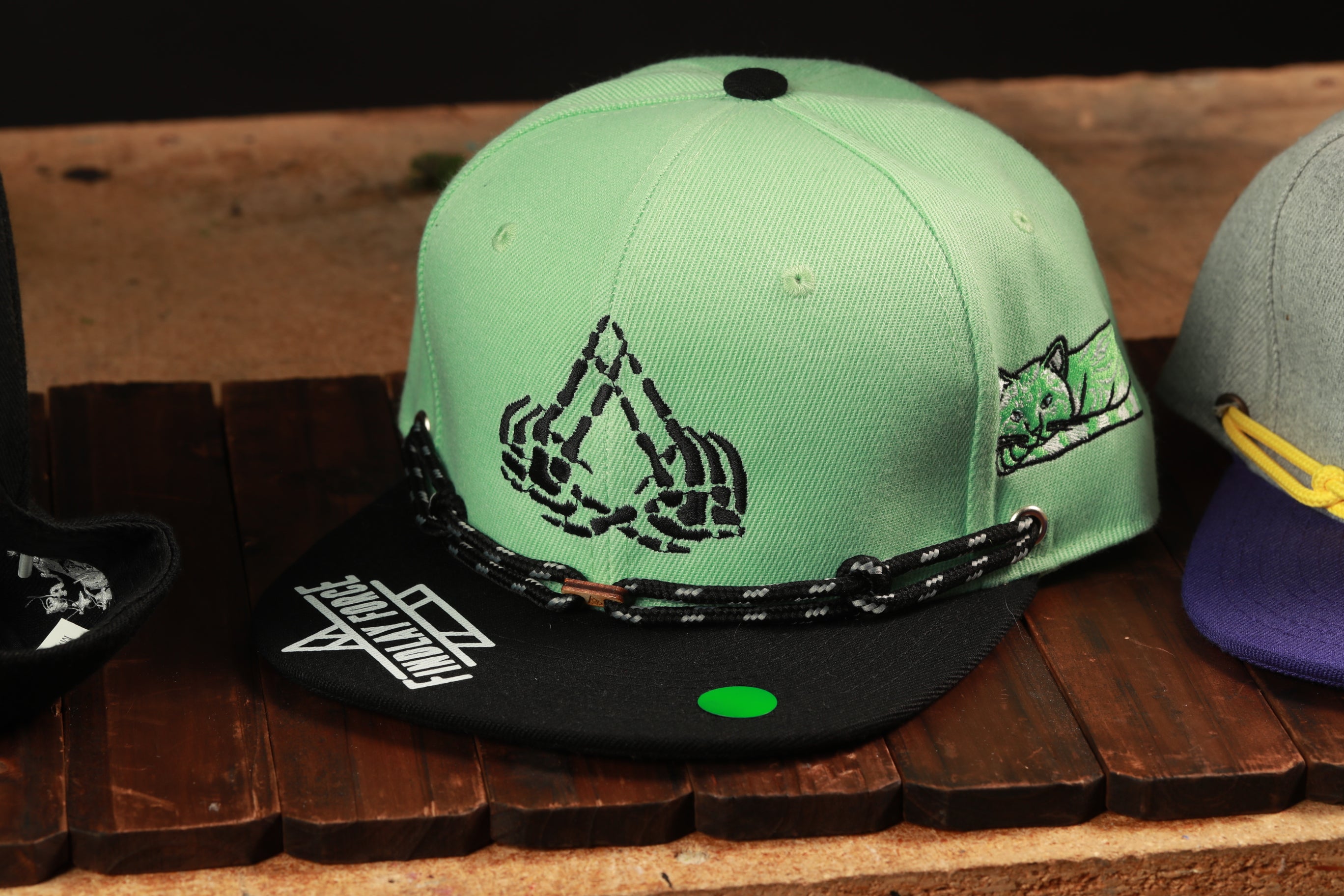 A green baseball cap with a skull and crossbones design, featuring a chain strap, is displayed on a wooden surface.