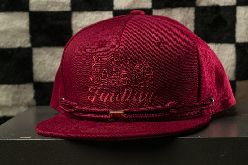 A burgundy baseball cap with a cat design and the word "Findlay" printed on it, sitting on a black surface against a checkered background.