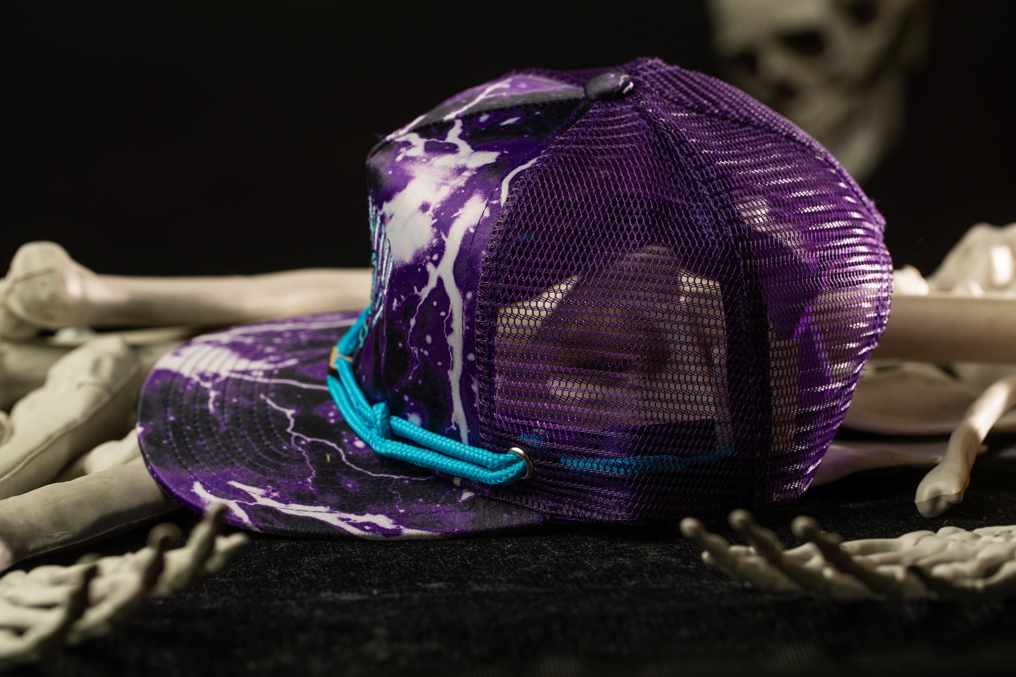 A purple mesh baseball cap with a blue strap is placed on a black surface, surrounded by skeletal bones.