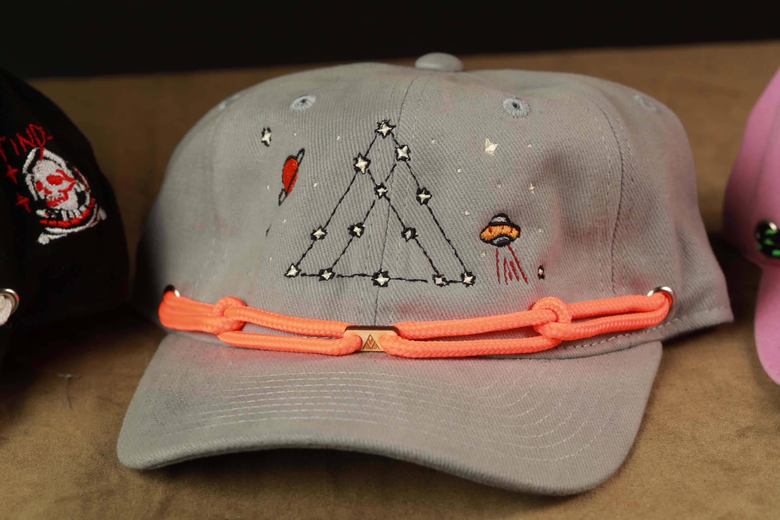 A gray baseball cap with a unique design featuring a triangle, stars, and an alien figure, along with an orange adjustable strap.