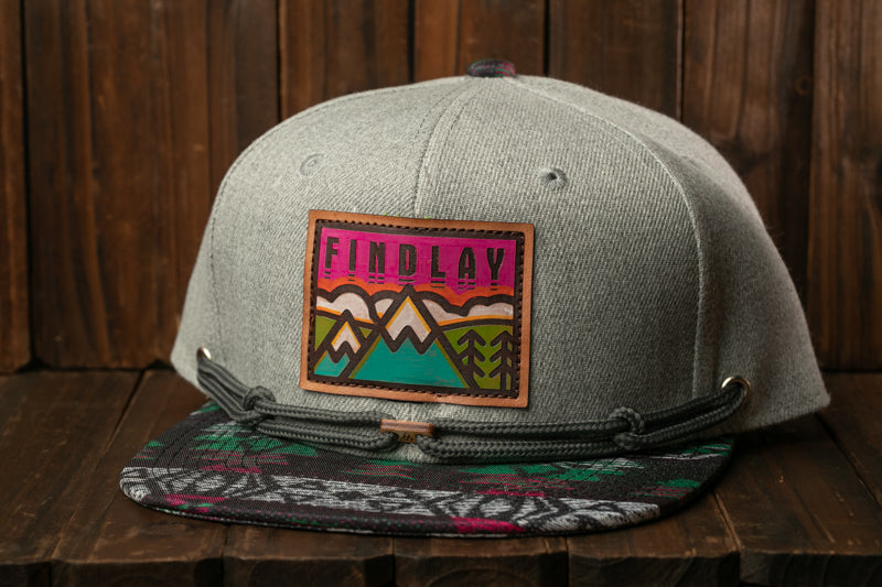 A gray baseball cap with a colorful patch on the front, featuring a mountain landscape design.