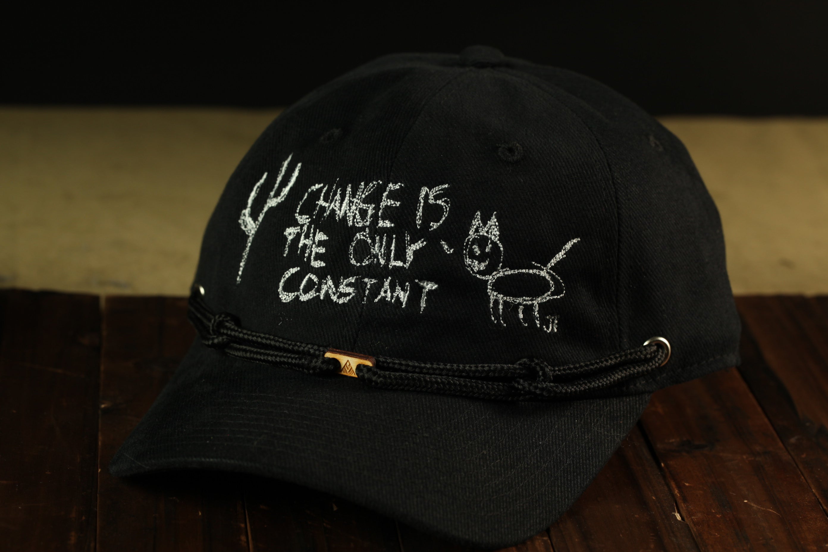 A black baseball cap with white text and a drawing on it, placed on a wooden surface.