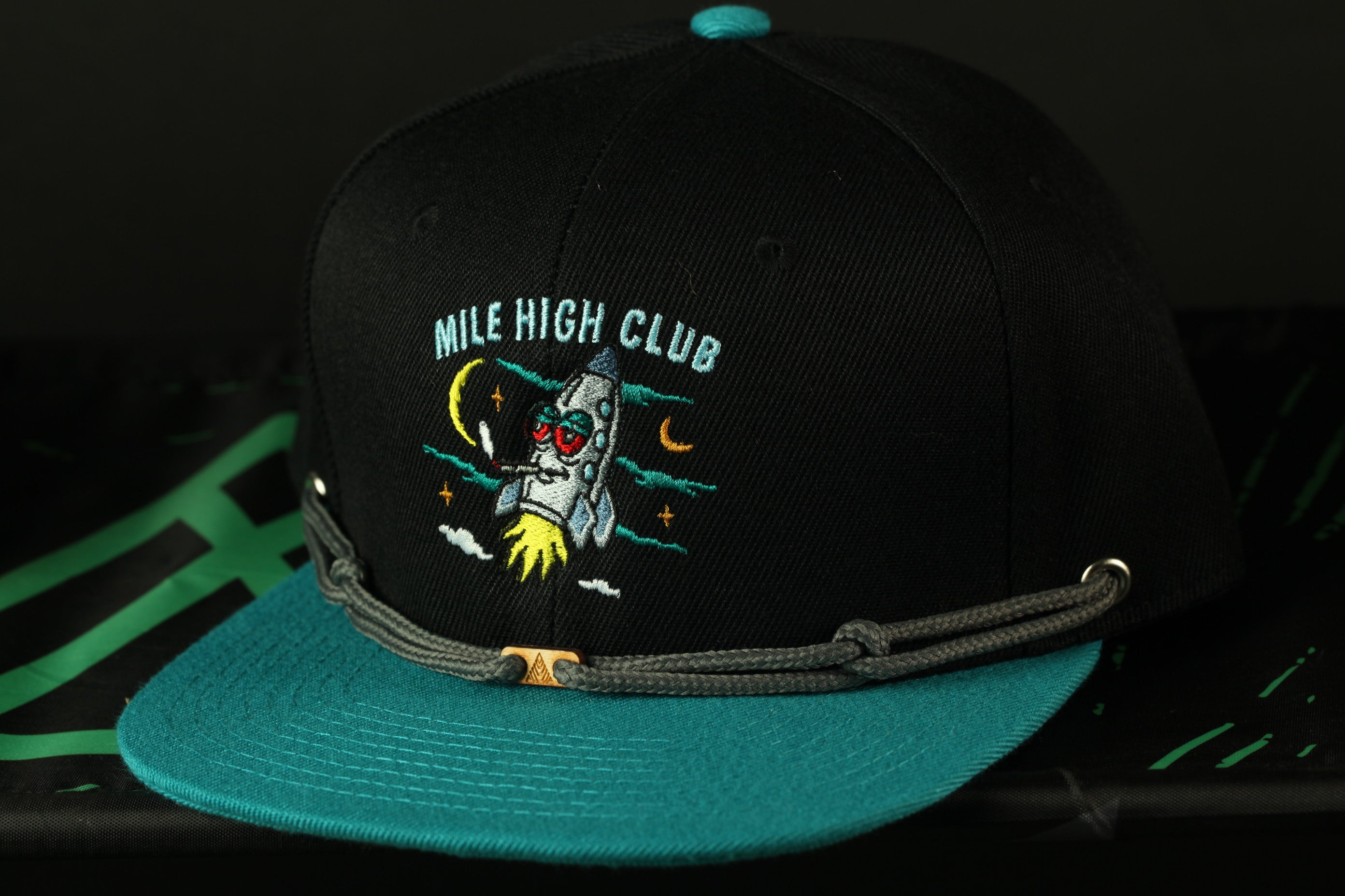 A black baseball cap with a teal brim and a graphic of a rocket ship on the front.