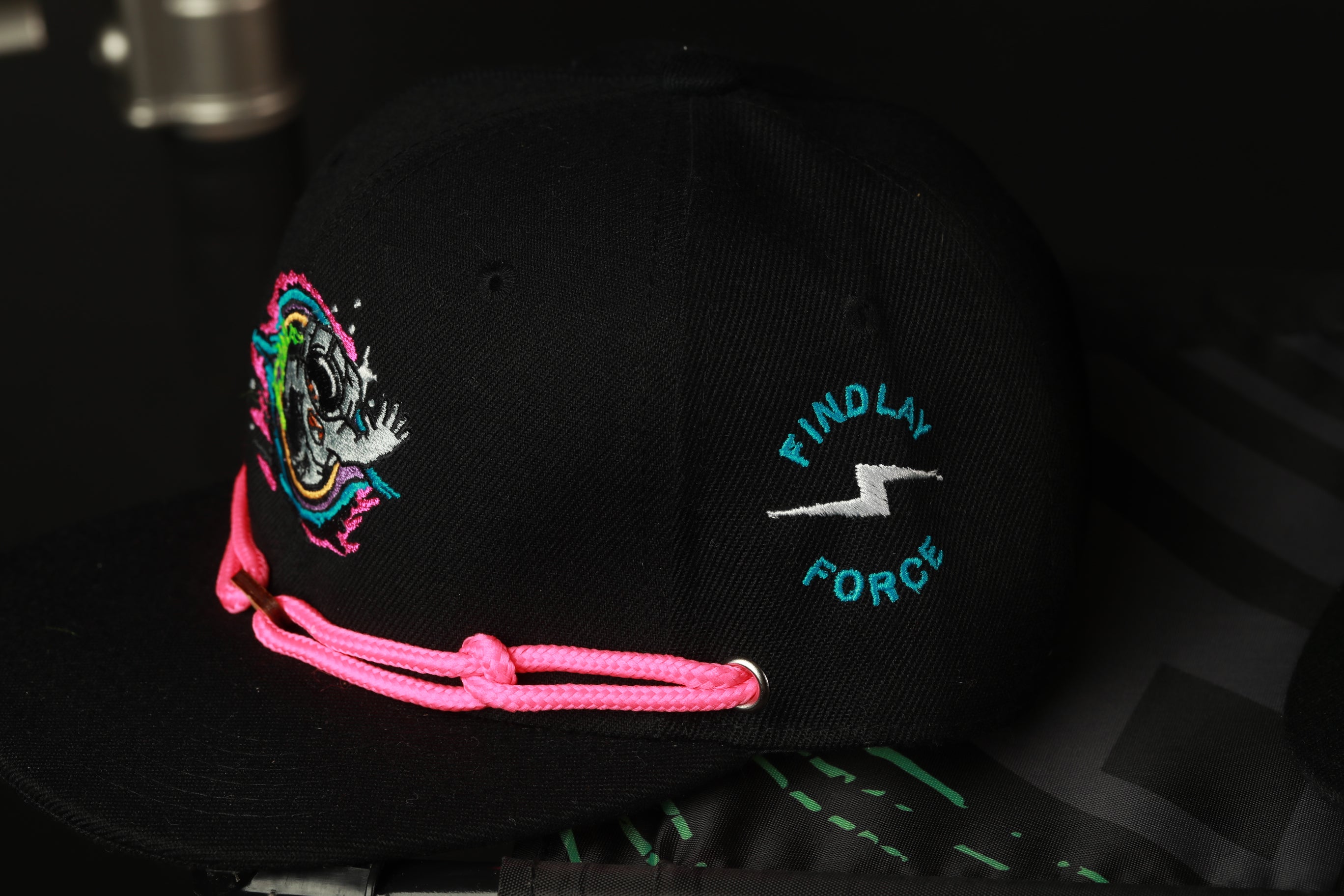 A black baseball cap with a pink string and a graphic design on the front.