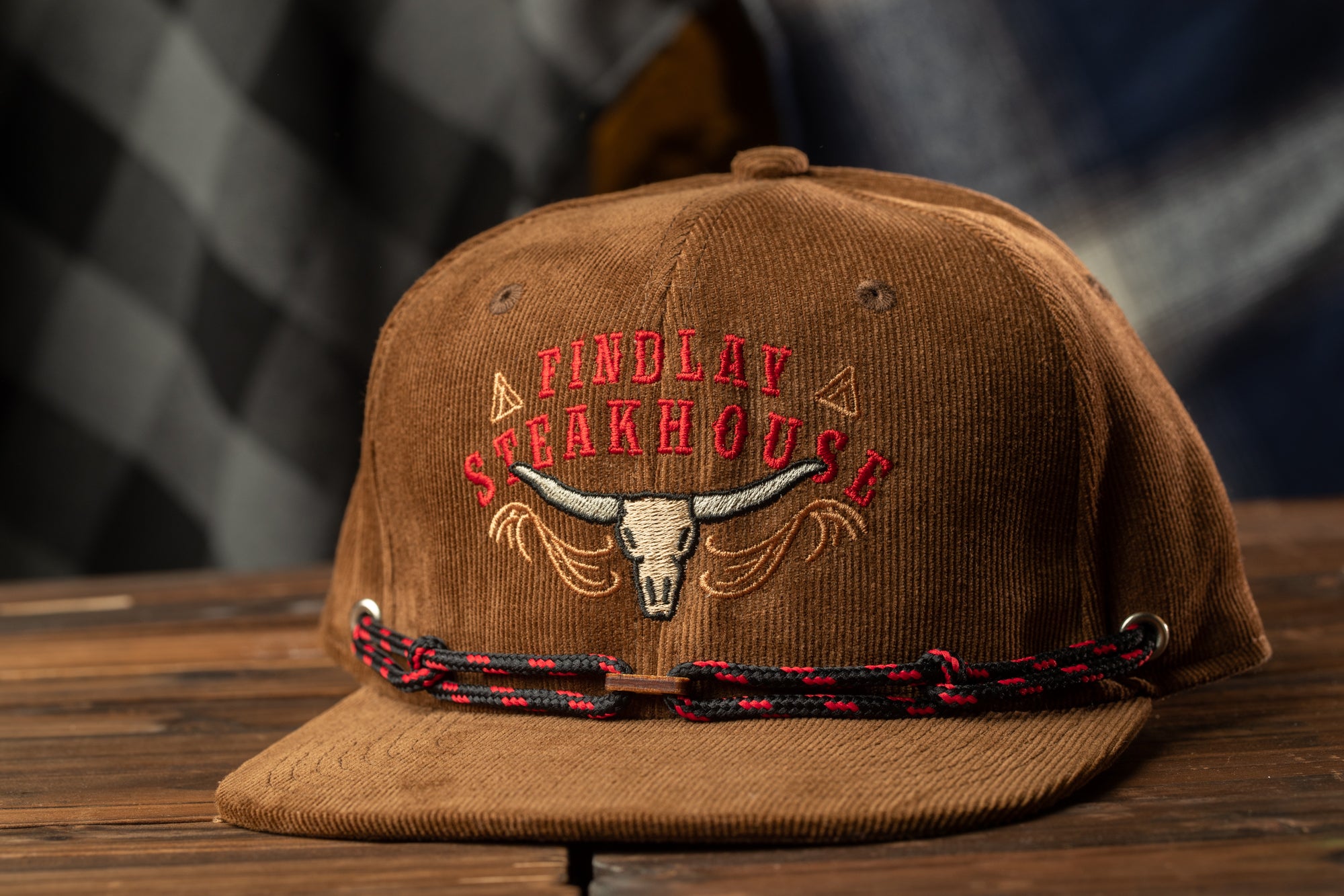 A brown baseball cap with the text "FINDlay Steakhouse" embroidered on it, sitting on a wooden surface.