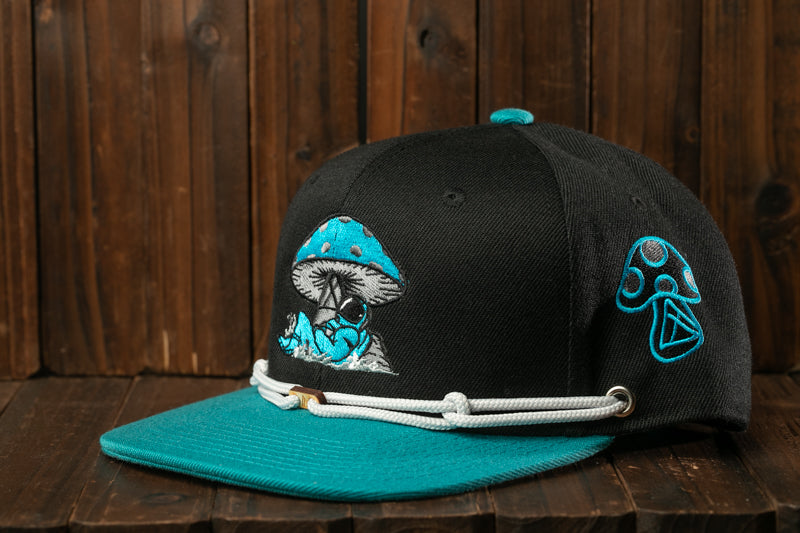 A black baseball cap with a teal brim and a mushroom design on the front, sitting on a wooden surface.