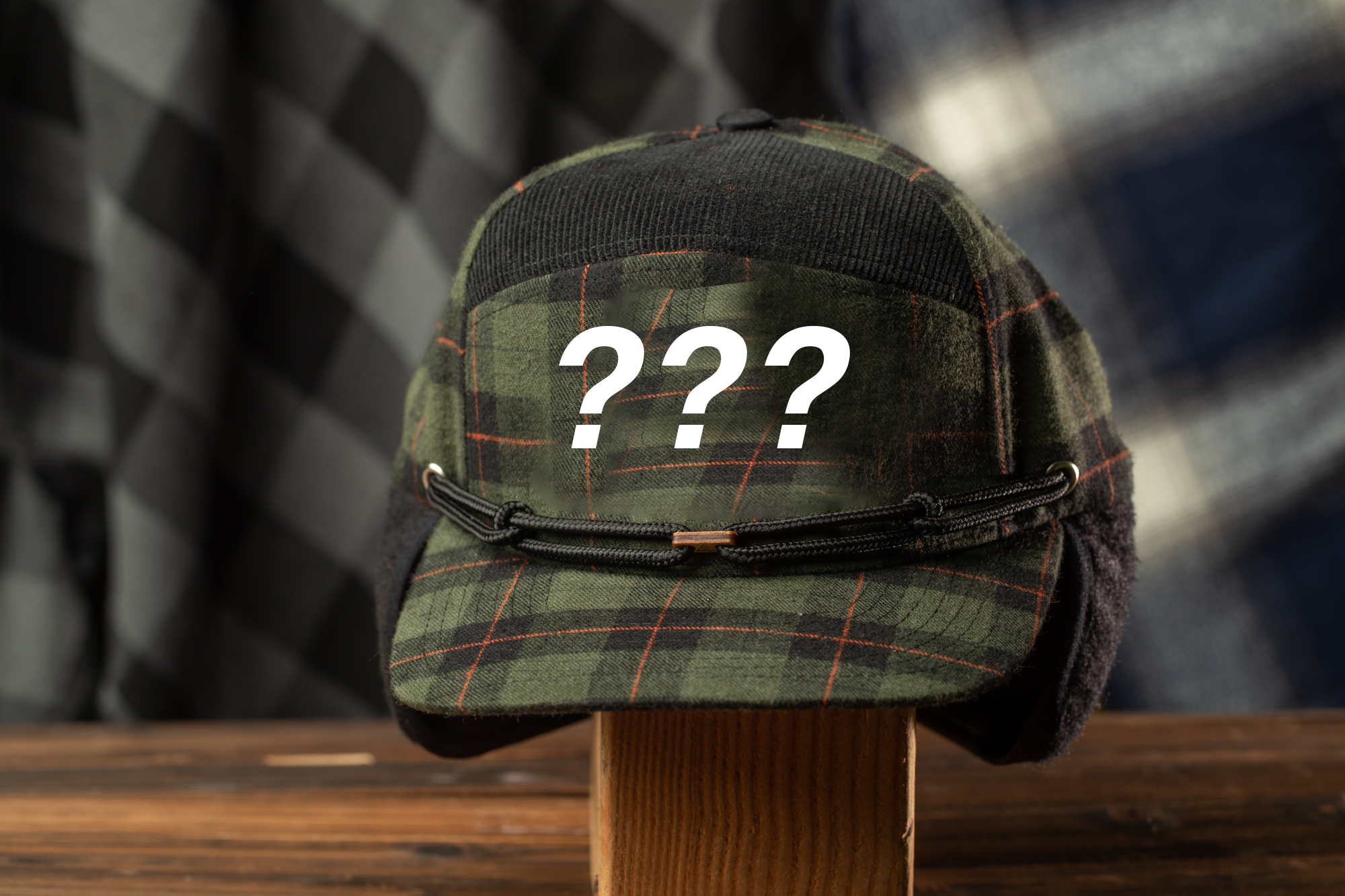 A plaid hat with a question mark symbol on it, displayed on a wooden stand against a checkered background.