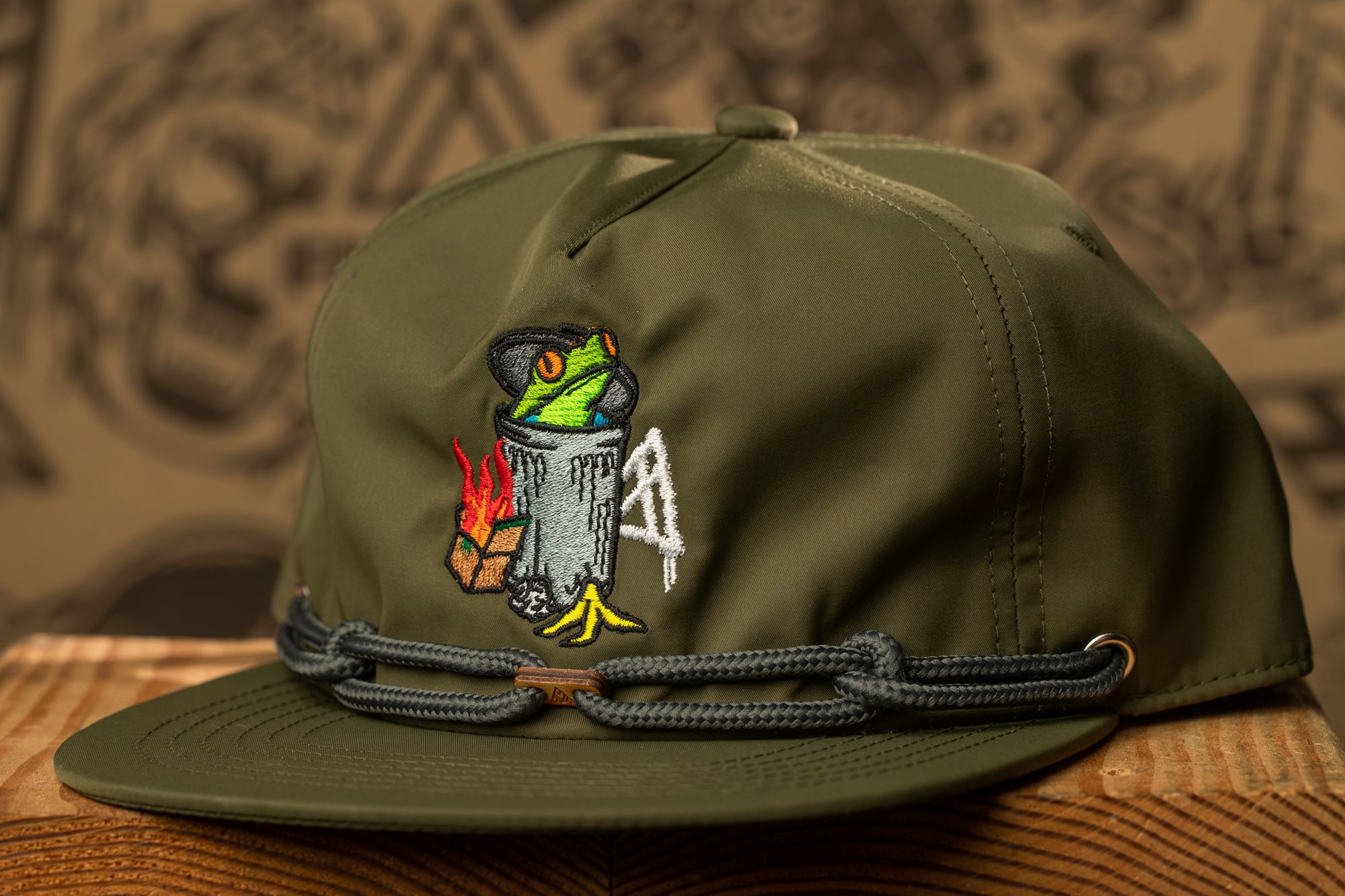 A green baseball cap with an embroidered design on the front, featuring a frog and a trash can.