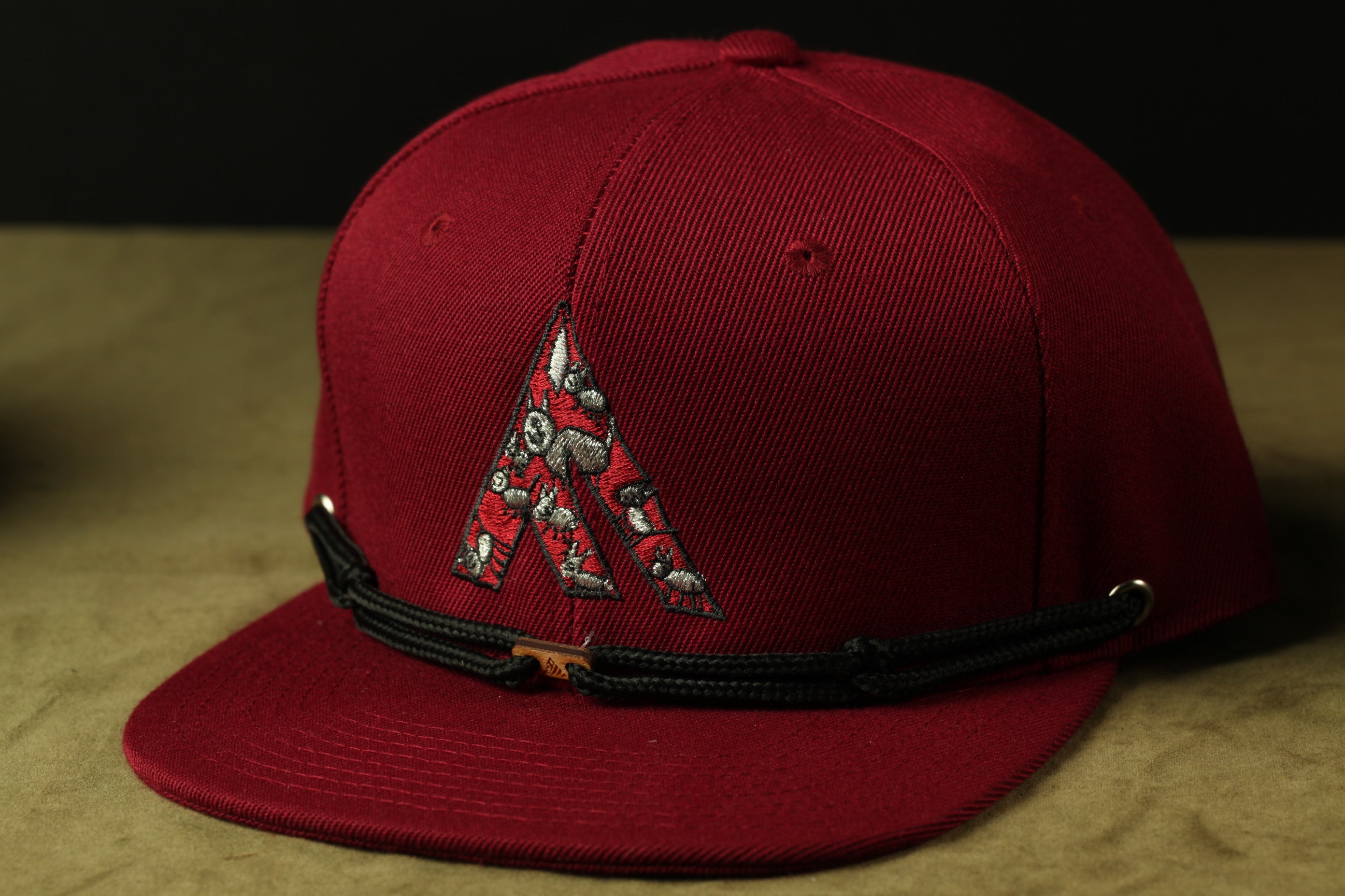 A maroon baseball cap with a black adjustable strap and a decorative logo on the front.