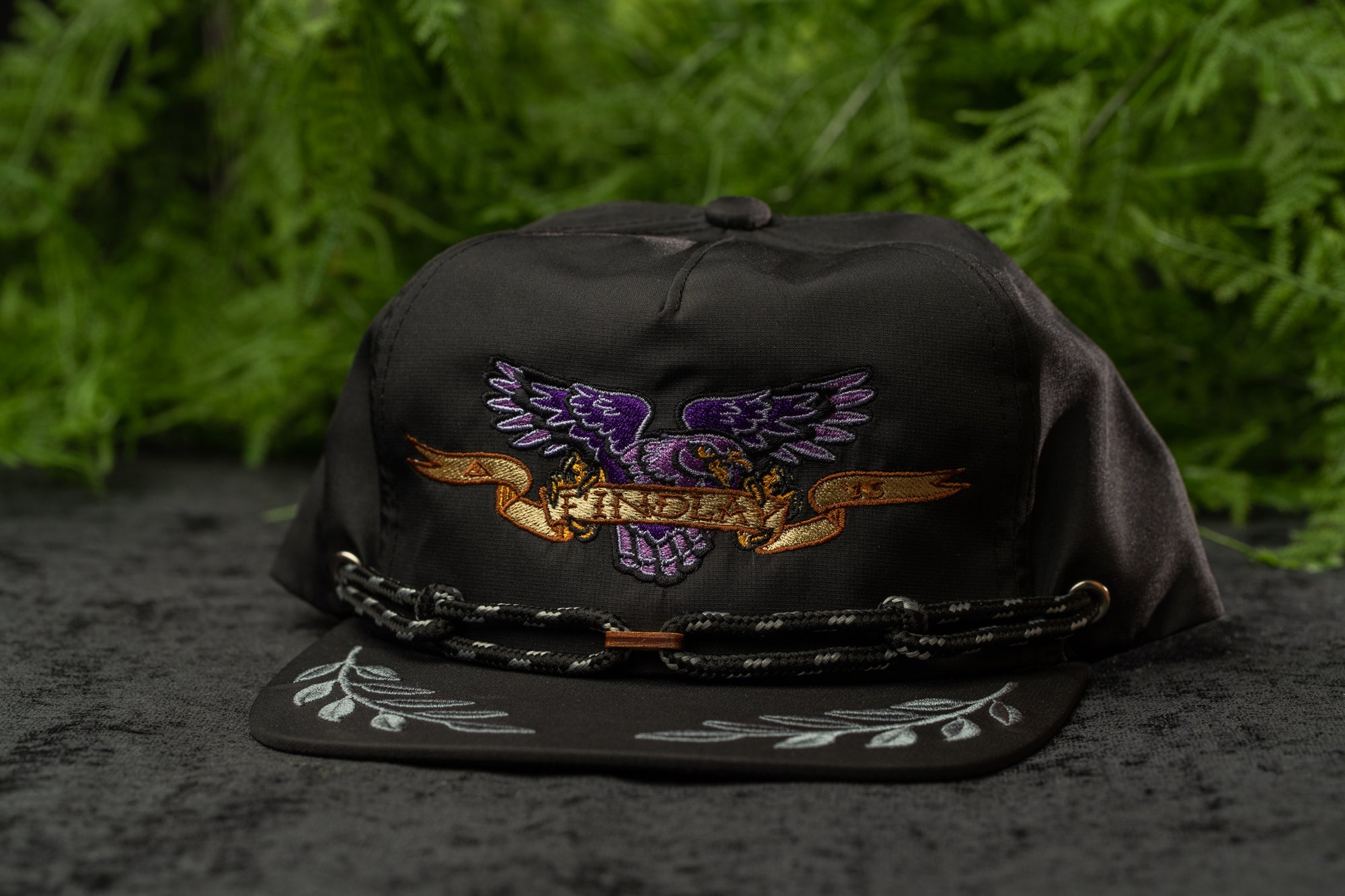 A black baseball cap with a purple eagle design and a banner that says "INDIA" on it, placed on a surface with green foliage in the background.