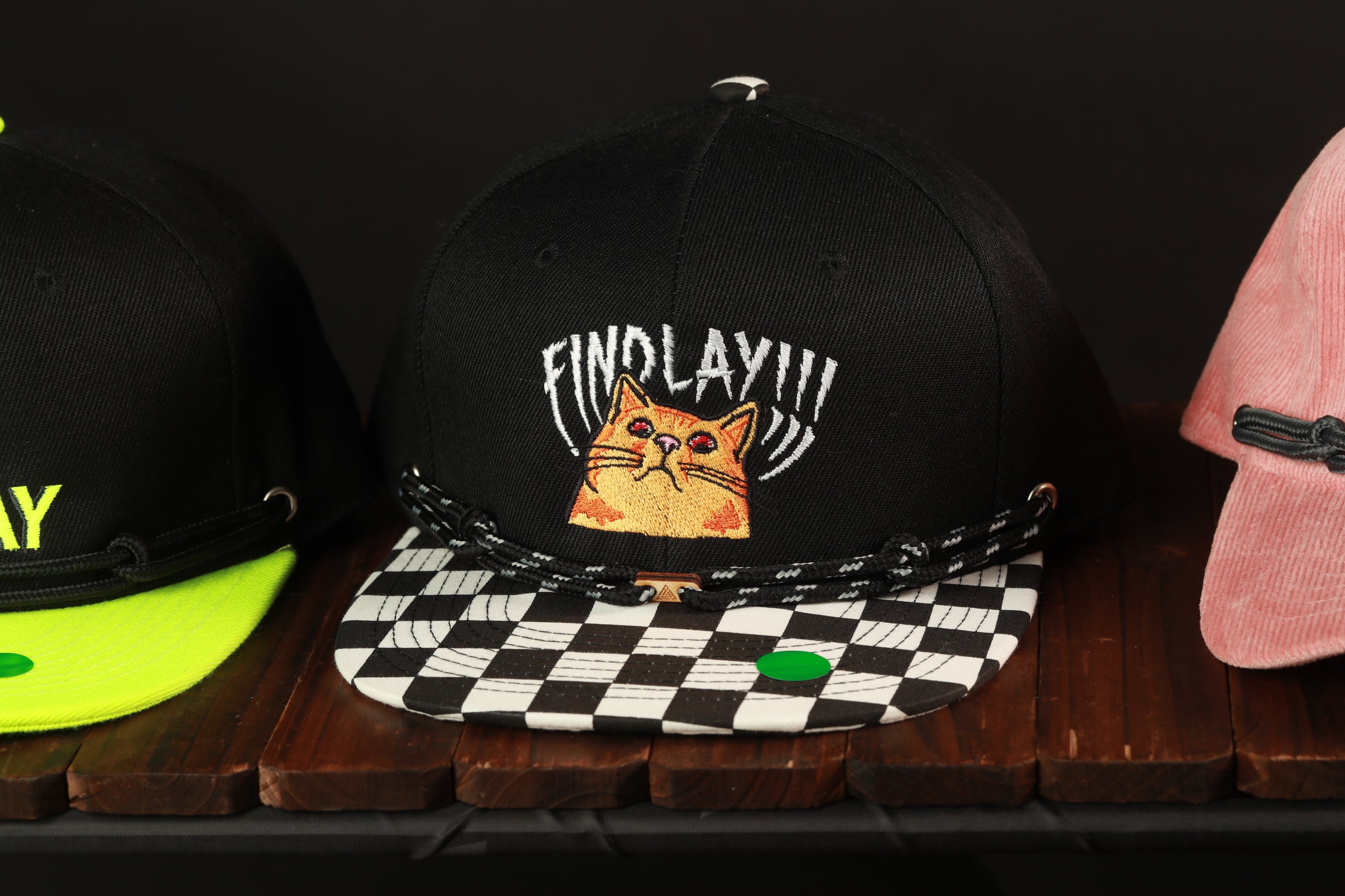 A black baseball cap with a cat embroidered on it, along with other hats, displayed on a wooden surface.