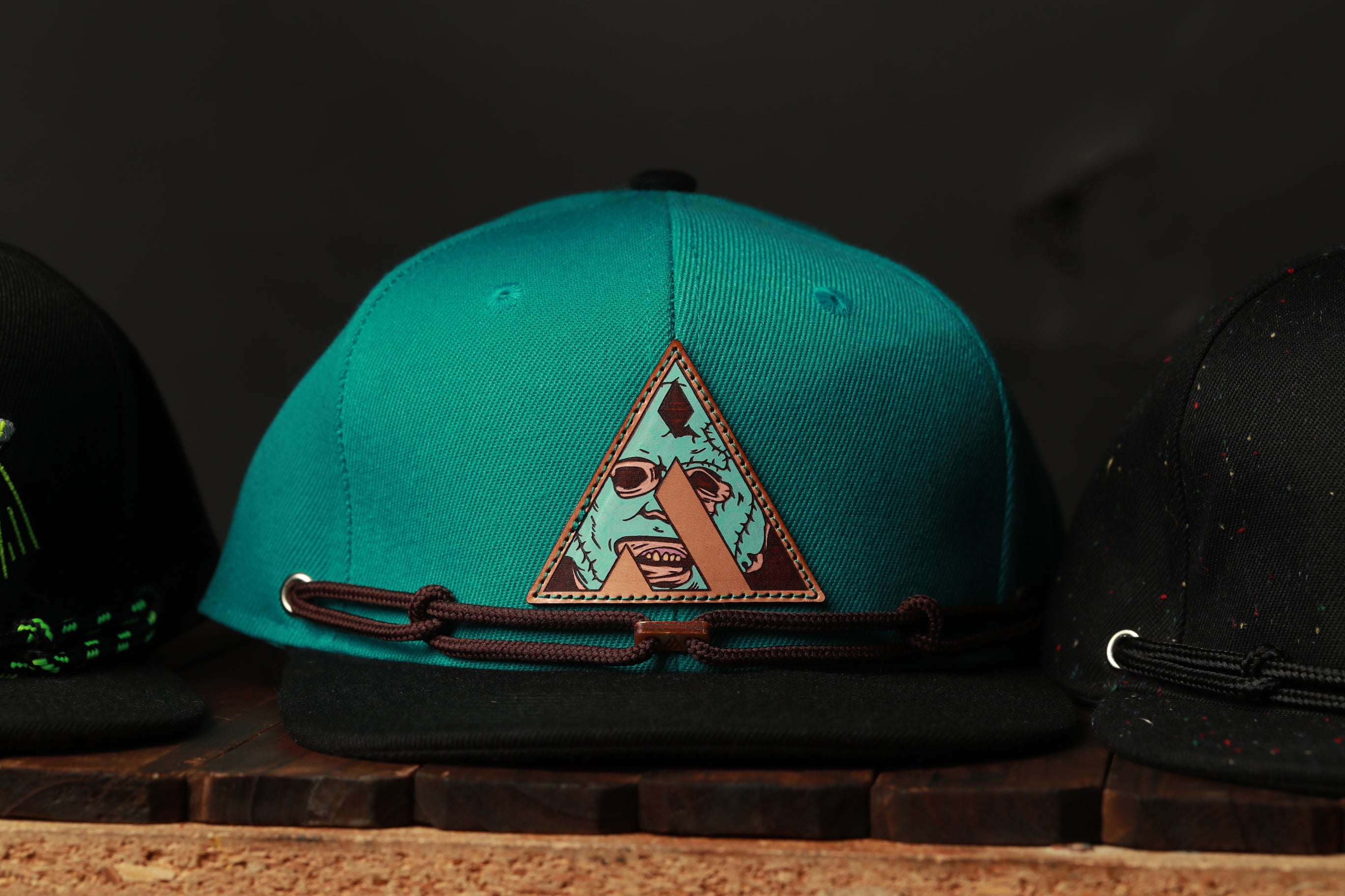 A turquoise baseball cap with a triangular logo featuring a face and a skull design, placed on a wooden surface.