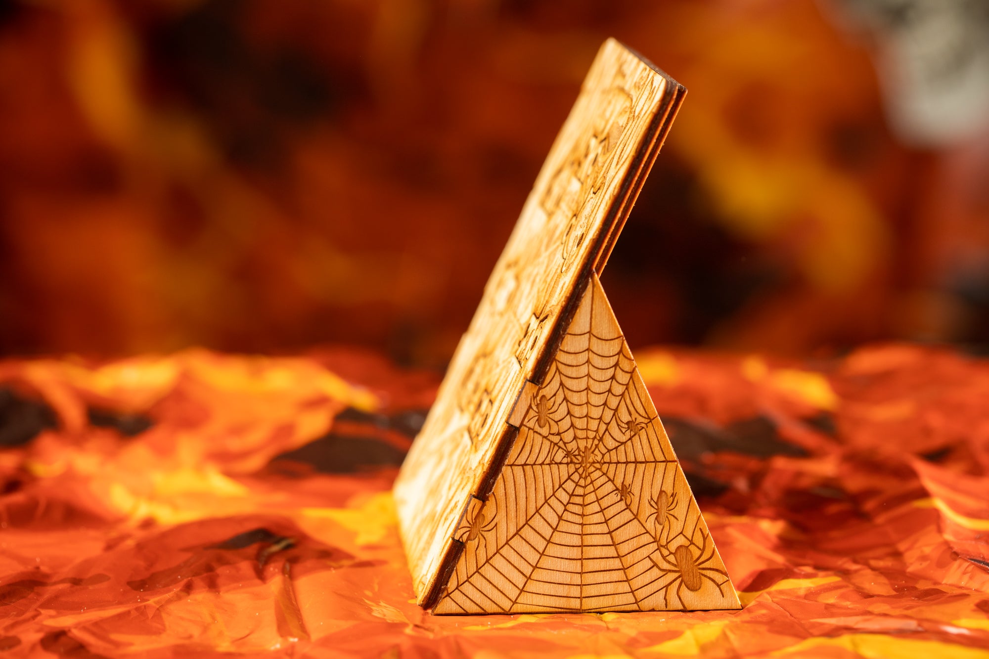 A wooden triangular object with a spider web pattern on it, placed on a surface with orange and yellow leaves.