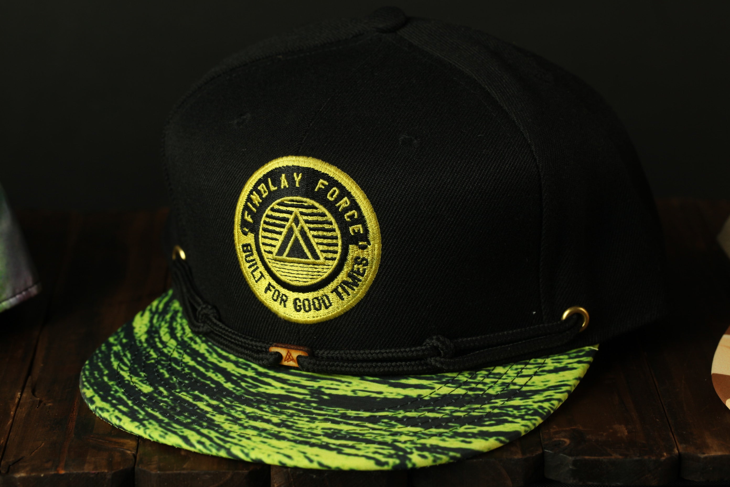 A black baseball cap with a yellow and green patterned brim, featuring a circular logo with text and a triangle design.