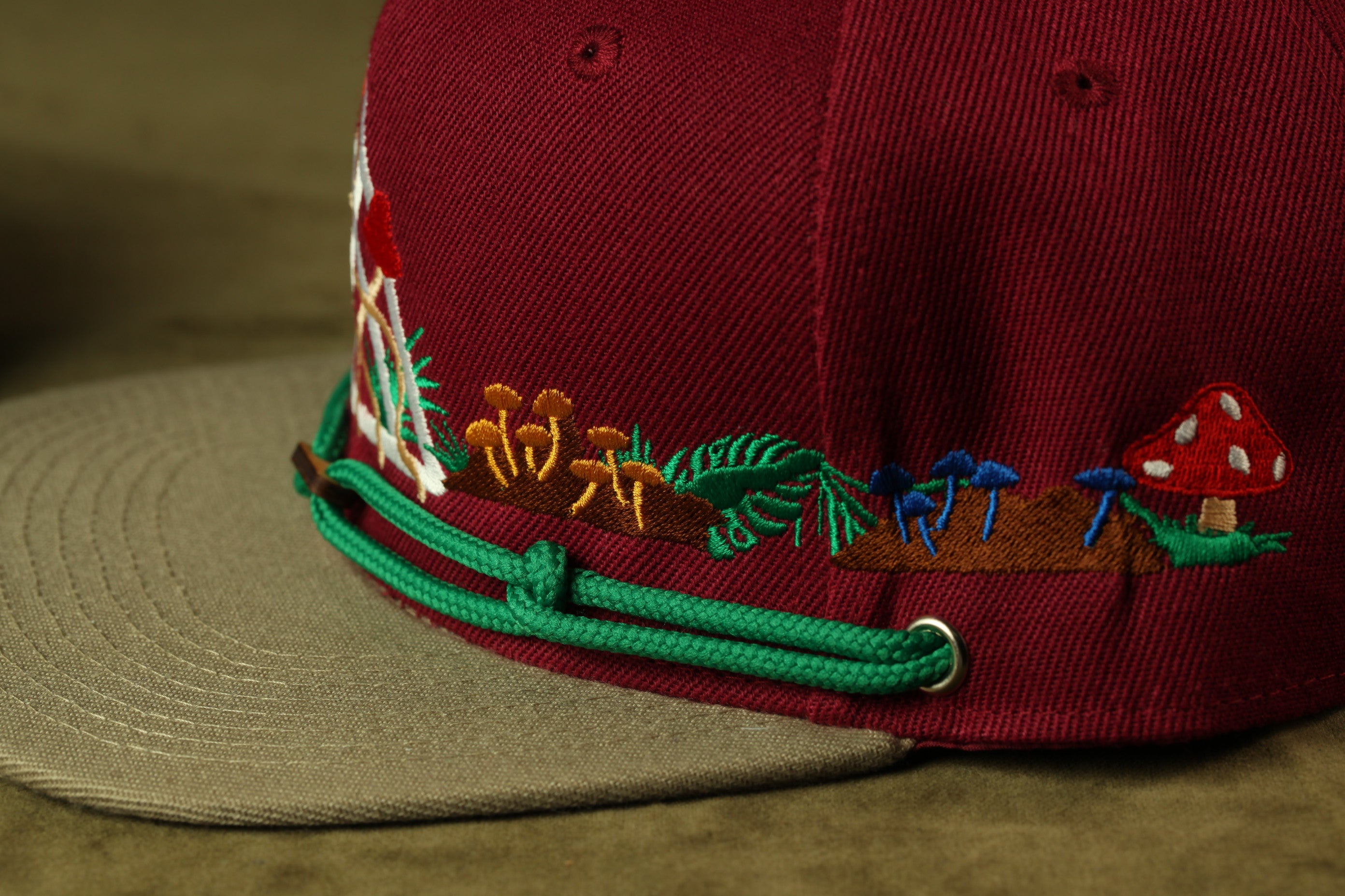 A close-up view of a burgundy baseball cap with colorful embroidered designs, including mushrooms, leaves, and other natural elements.