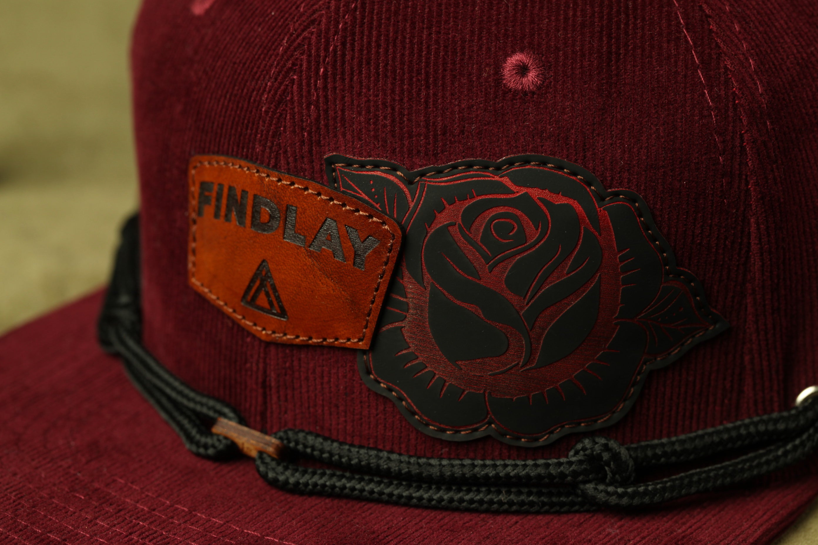 A close-up view of a burgundy baseball cap with a black rose embroidered on the front and a leather patch with the word "FINDlay" printed on it.