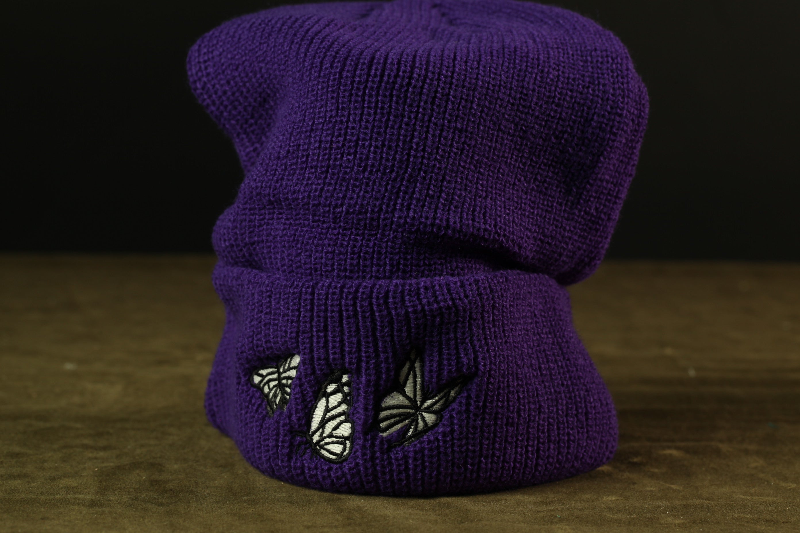 A purple knit beanie with a butterfly design on the front is shown against a dark background.