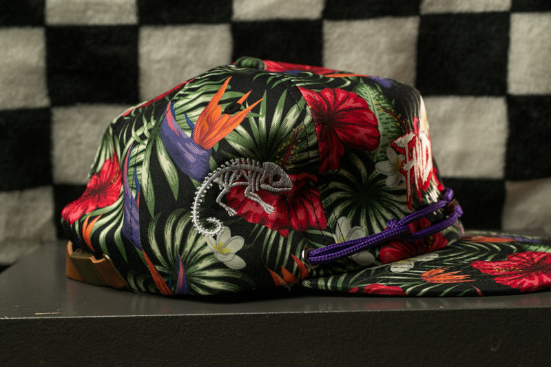 A colorful floral cap with a purple string and a small chameleon design on it, placed on a black surface against a checkered background.