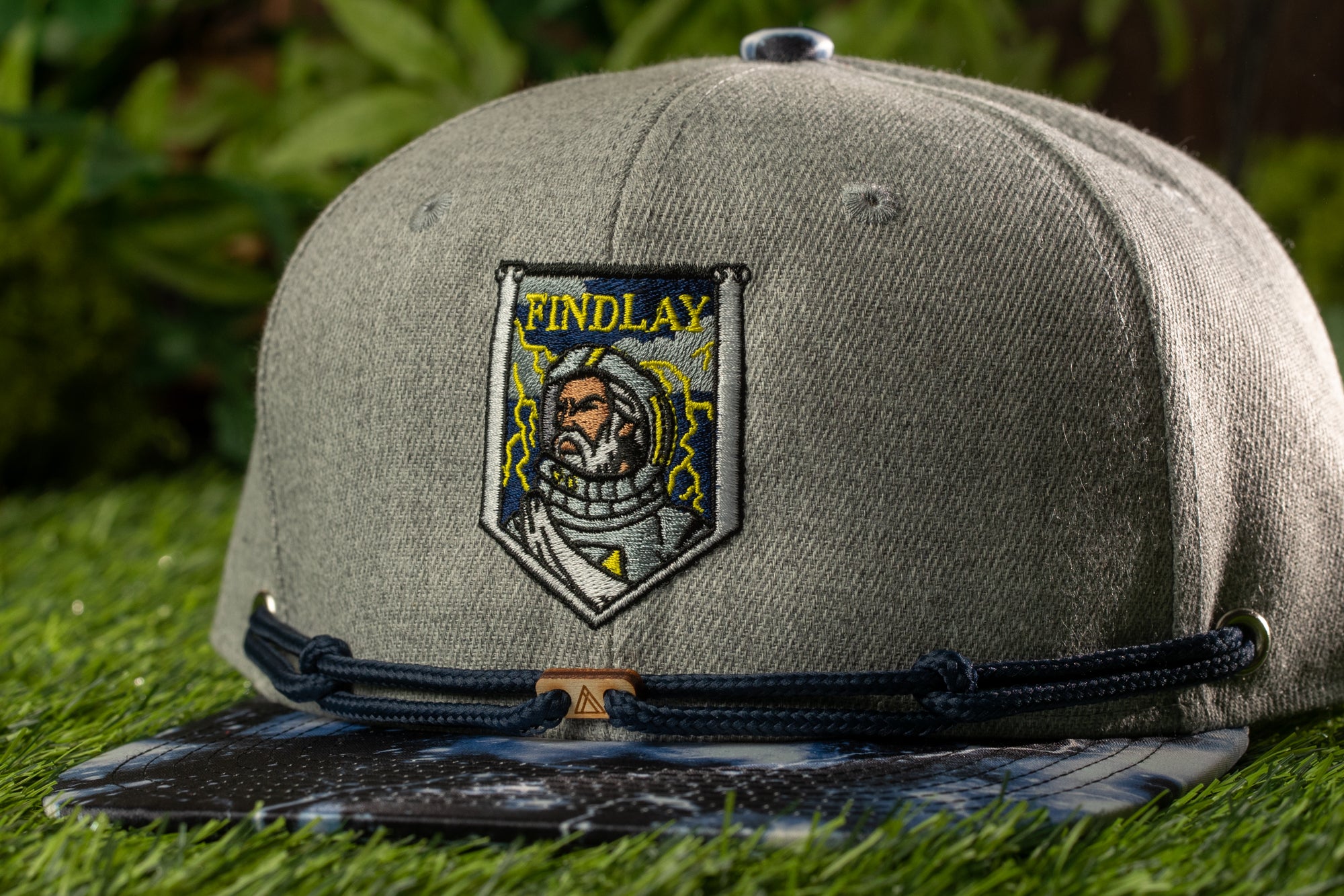 A gray baseball cap with a blue and white patterned brim, featuring a patch with the word "FINDLAY" and an illustration of a person in a spacesuit.