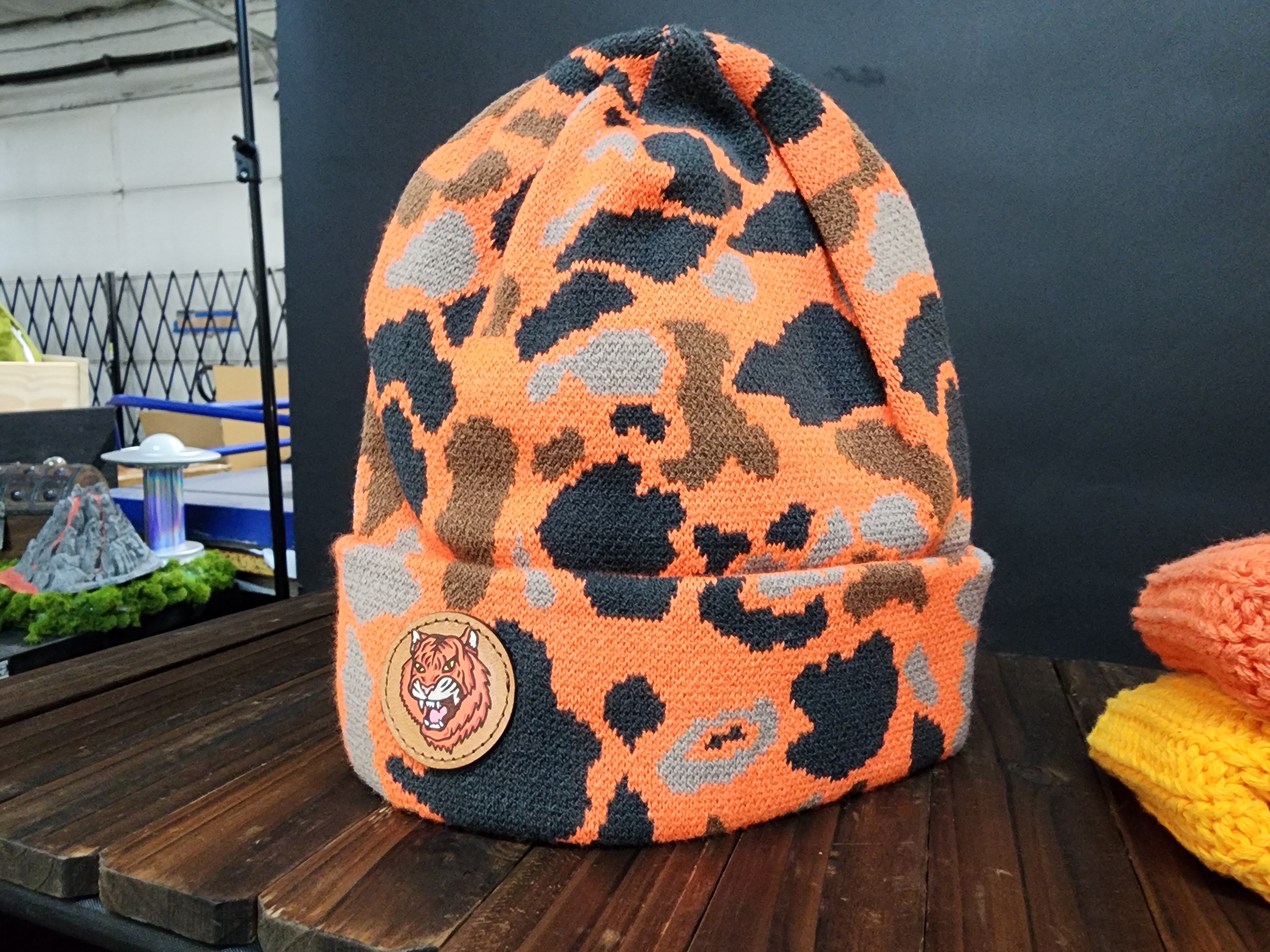 A camouflage-patterned orange and black beanie with a tiger logo is displayed on a wooden surface.