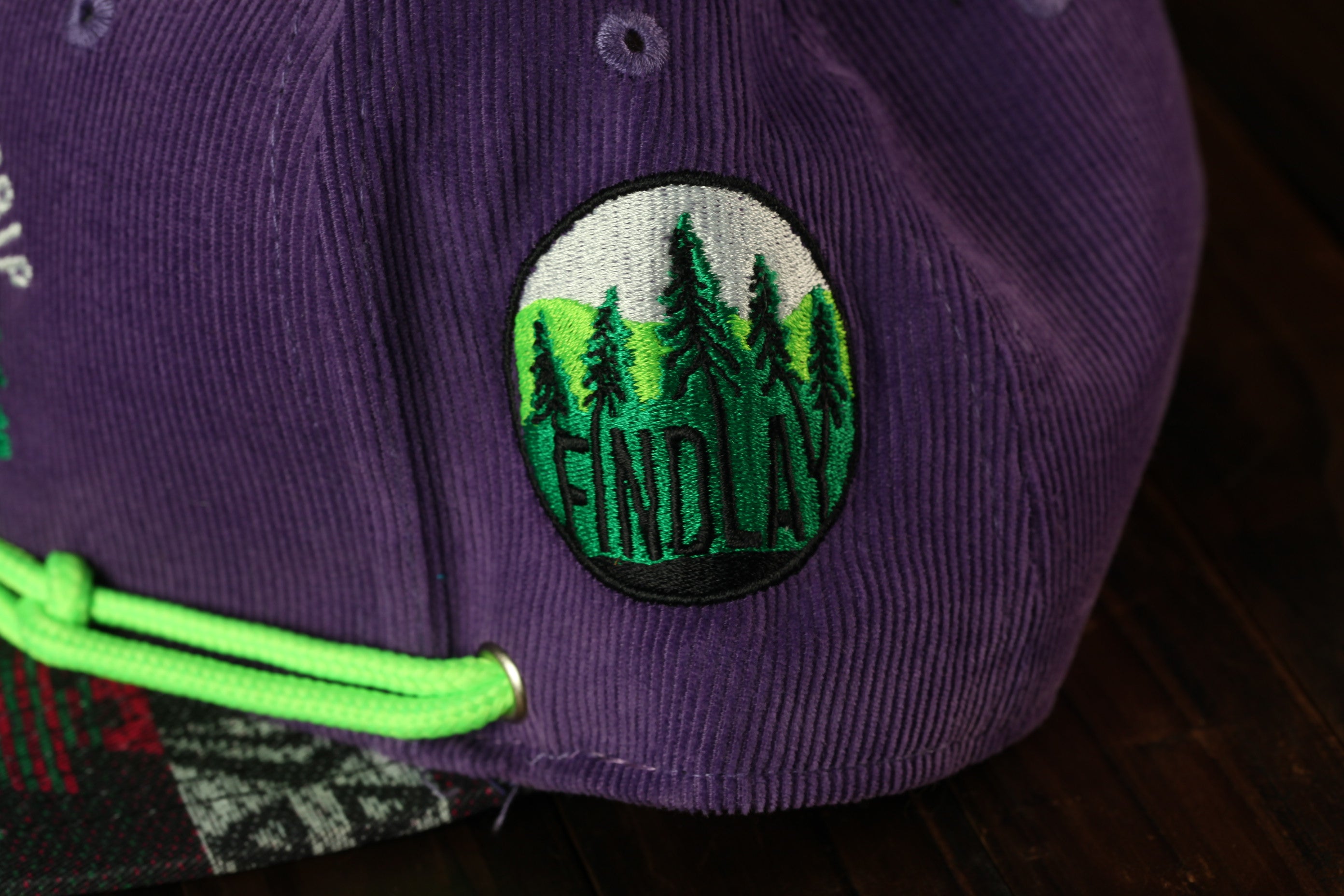 A close-up view of a purple baseball cap with a green string and a circular embroidered patch on the front.