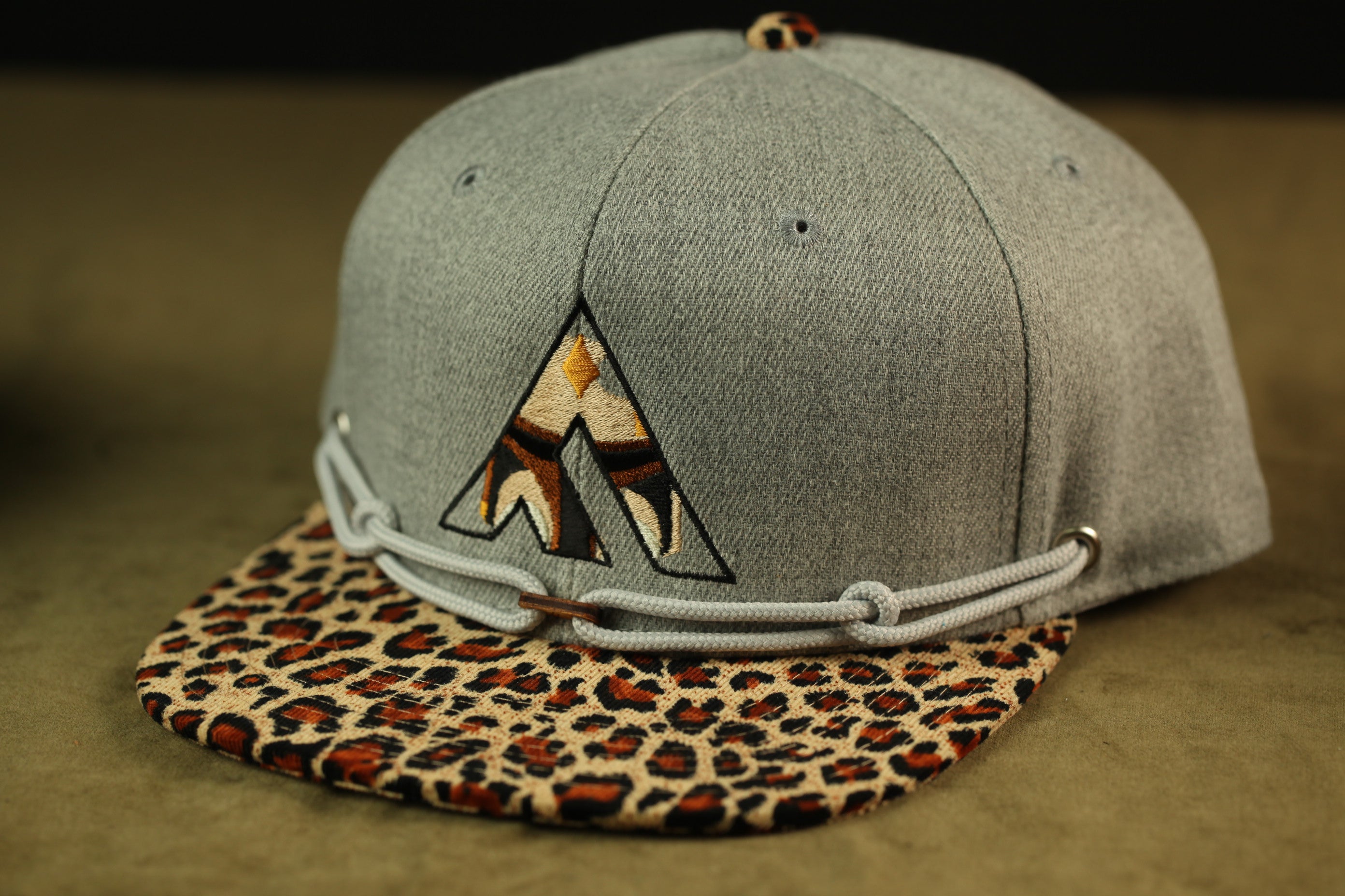 A gray baseball cap with a leopard print pattern and a geometric logo on the front.