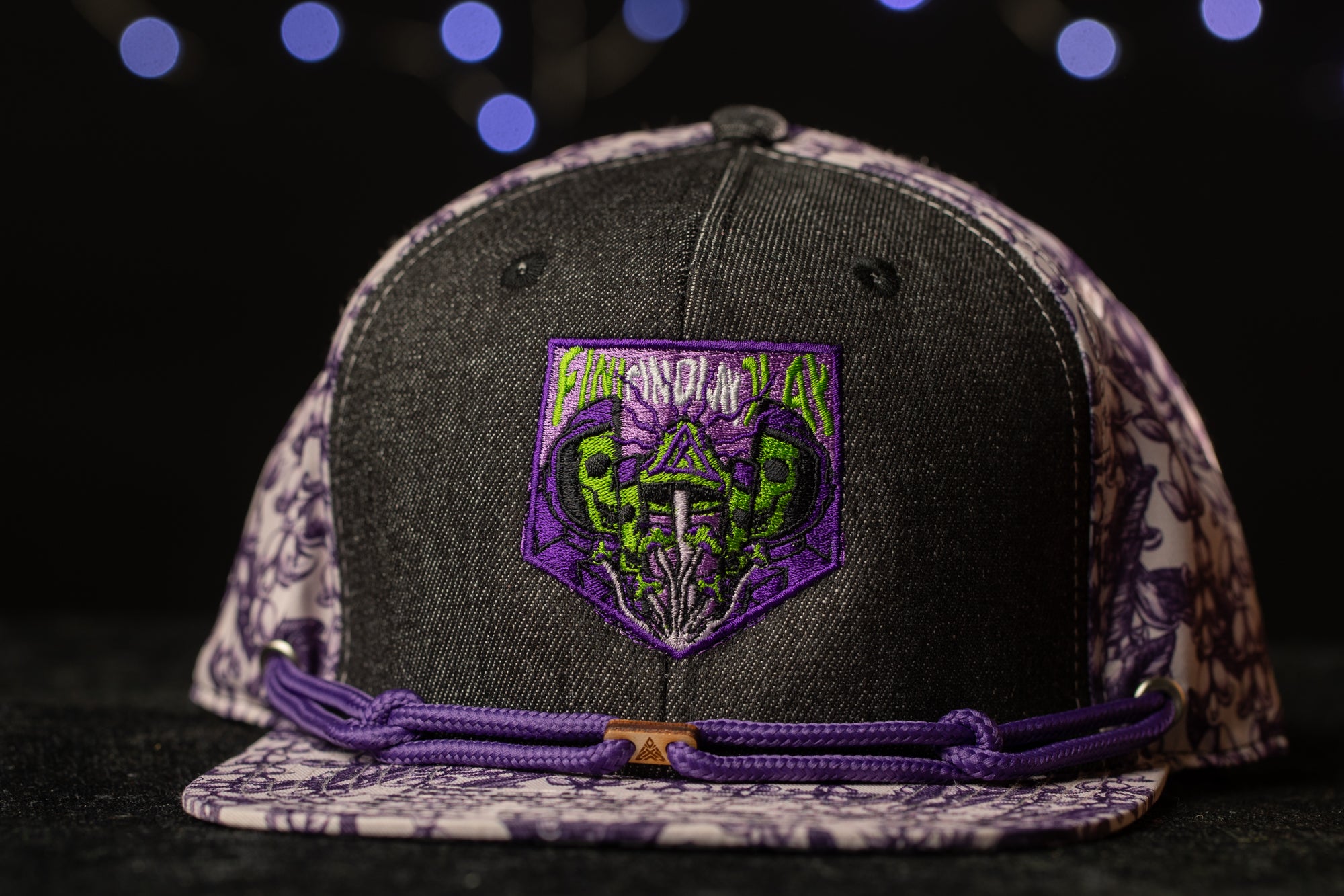 A black baseball cap with a purple and white floral pattern, featuring a purple patch with a skull and crossbones design.