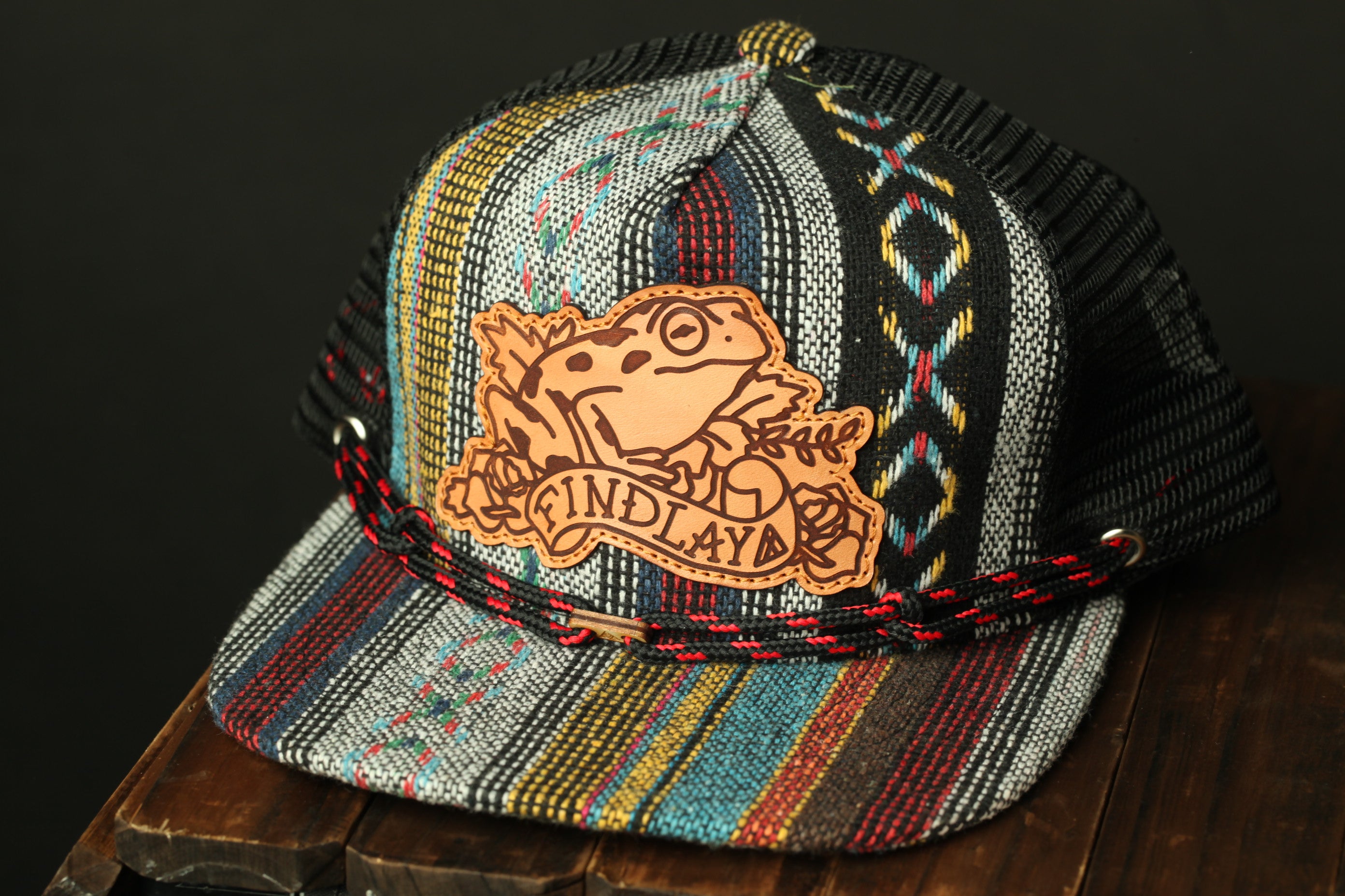 A colorful, patterned baseball cap with a leather frog-shaped patch on the front.