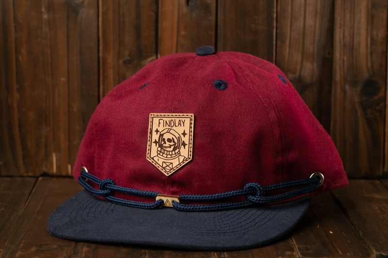A maroon baseball cap with a blue brim and a brown leather patch on the front, featuring a skull and crossbones design.