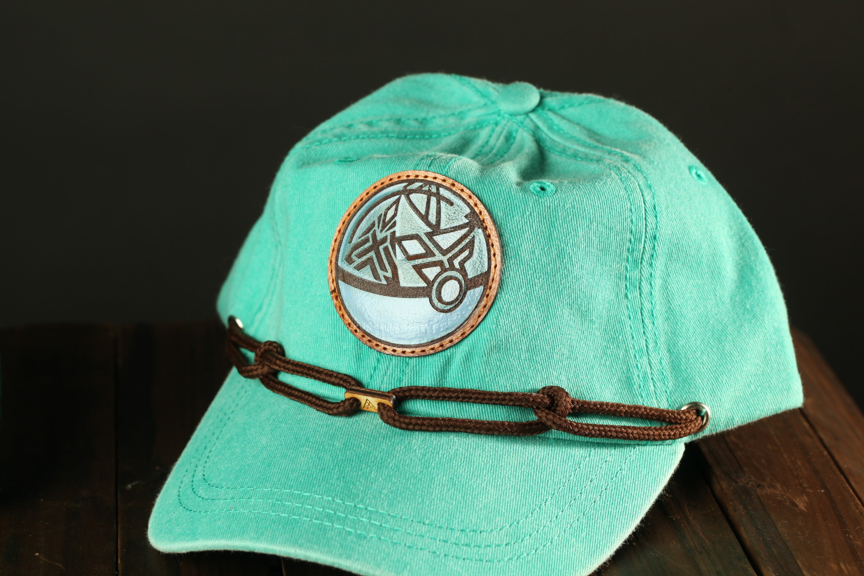 A turquoise baseball cap with a circular patch on the front and a brown chain strap.